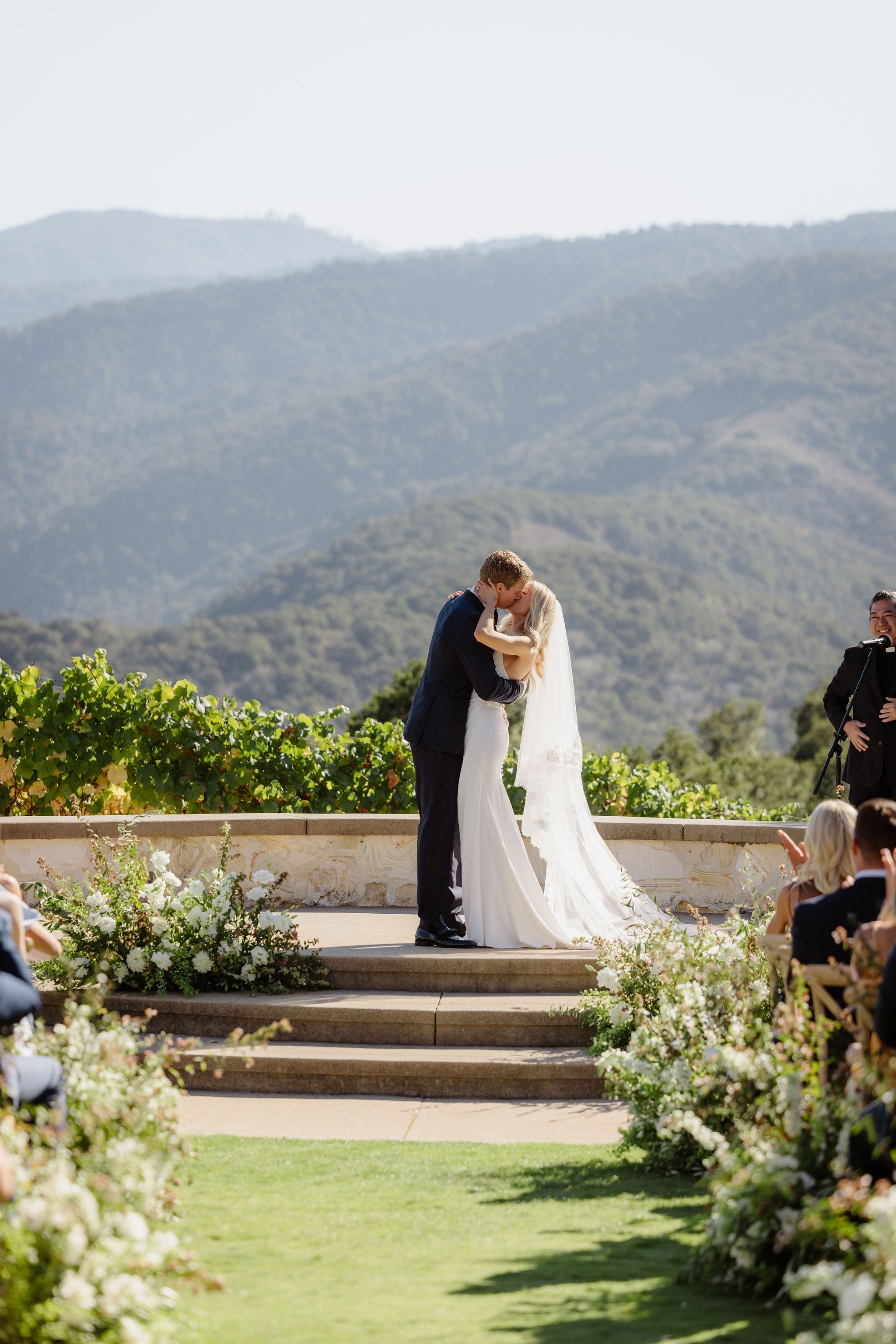 A bride and groom share a kiss during their outdoor wedding ceremony on steps surrounded by greenery and flowers, with mountainous landscape in the background and guests seated nearby.
