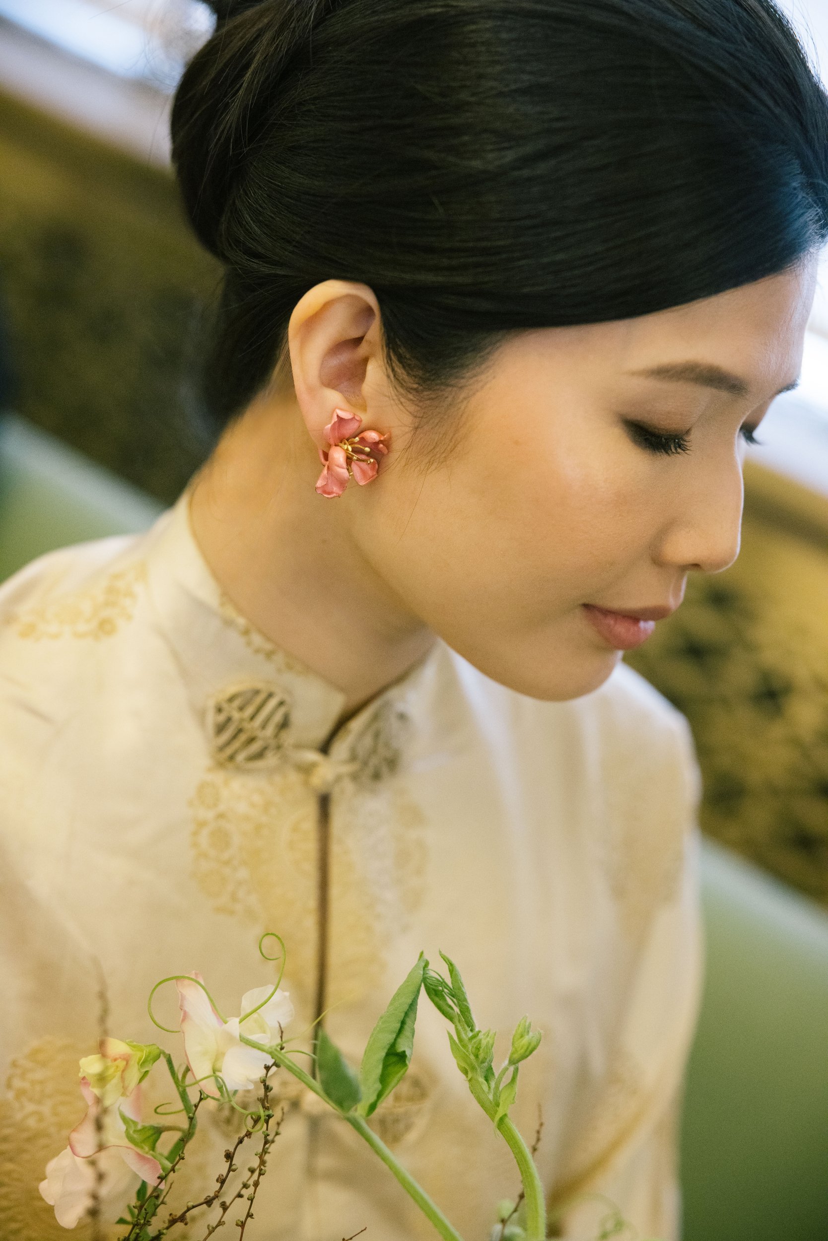 A woman with black hair styled elegantly, wearing floral earrings and traditional clothing, appears to be looking down at flowers. The background is blurred with a soft, natural setting.