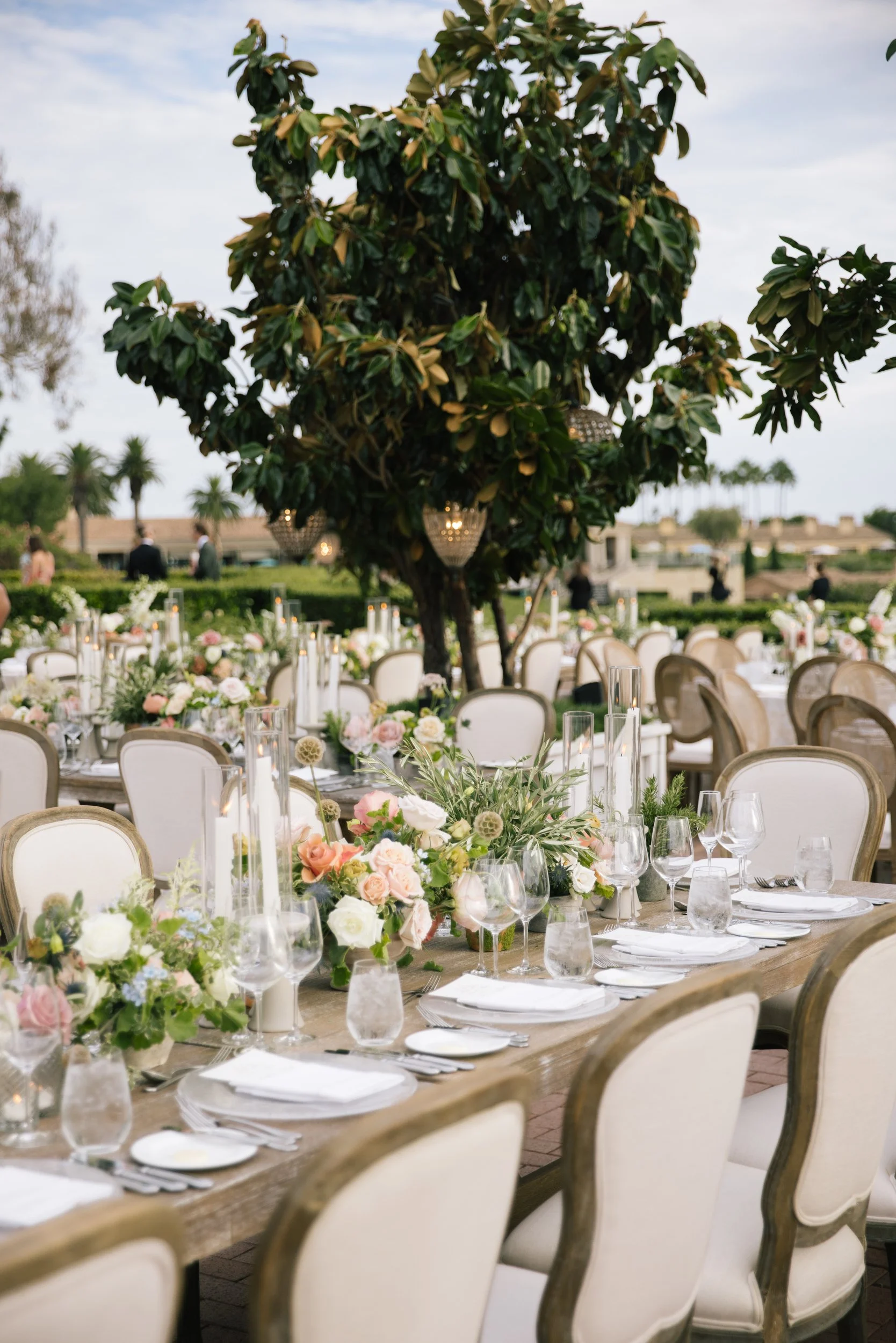 A large outdoor wedding reception setup featuring a long wooden table decorated with floral arrangements in shades of pink, white, and green. Elegant glassware and white plates are arranged on the table, with cream-colored chairs surrounding it. In t