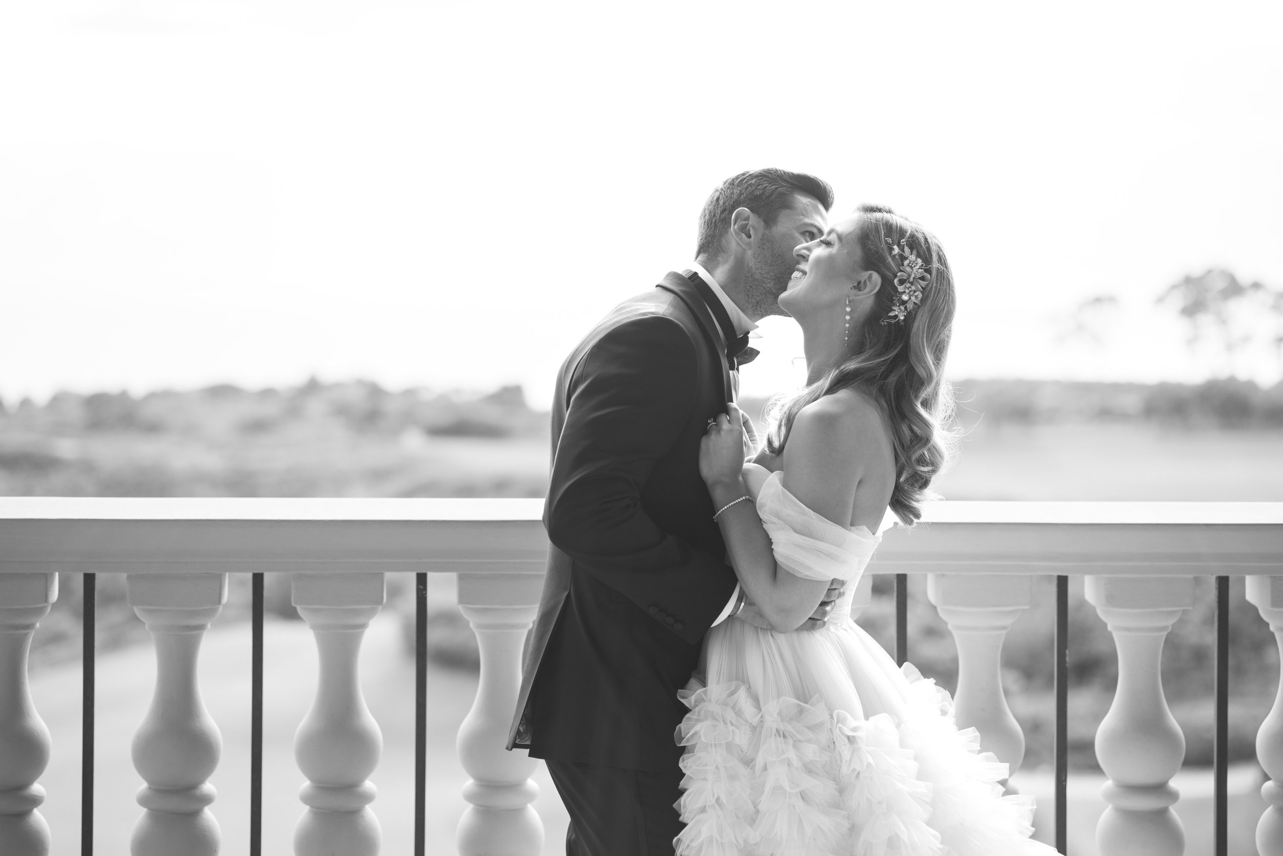 A black and white photo of a bride and groom on a balcony, embracing and about to kiss, outdoors with a landscape in the background.