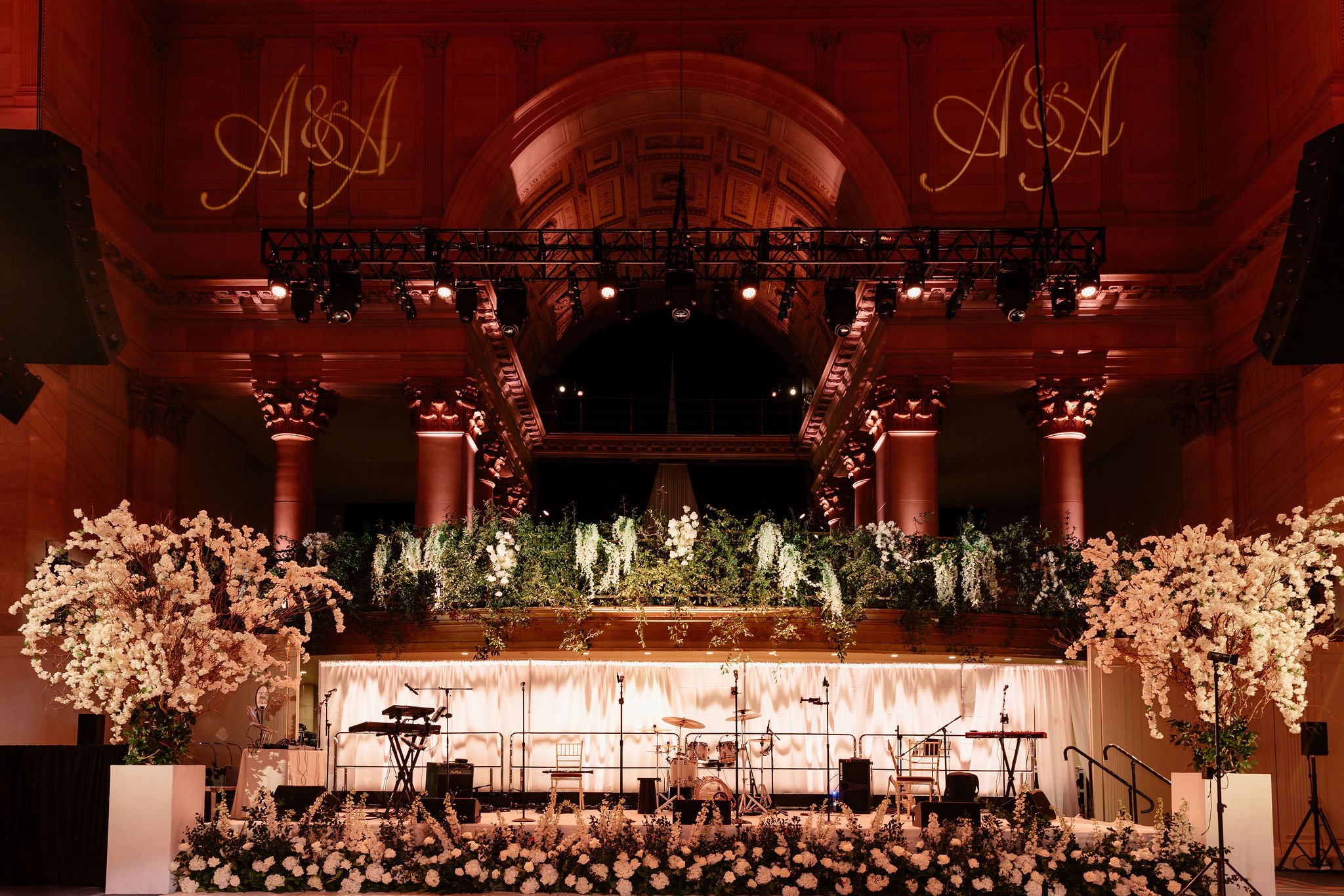 Indoor concert venue decorated with flowers and stage equipment, large illuminated initials 'A & M' projected on the back wall