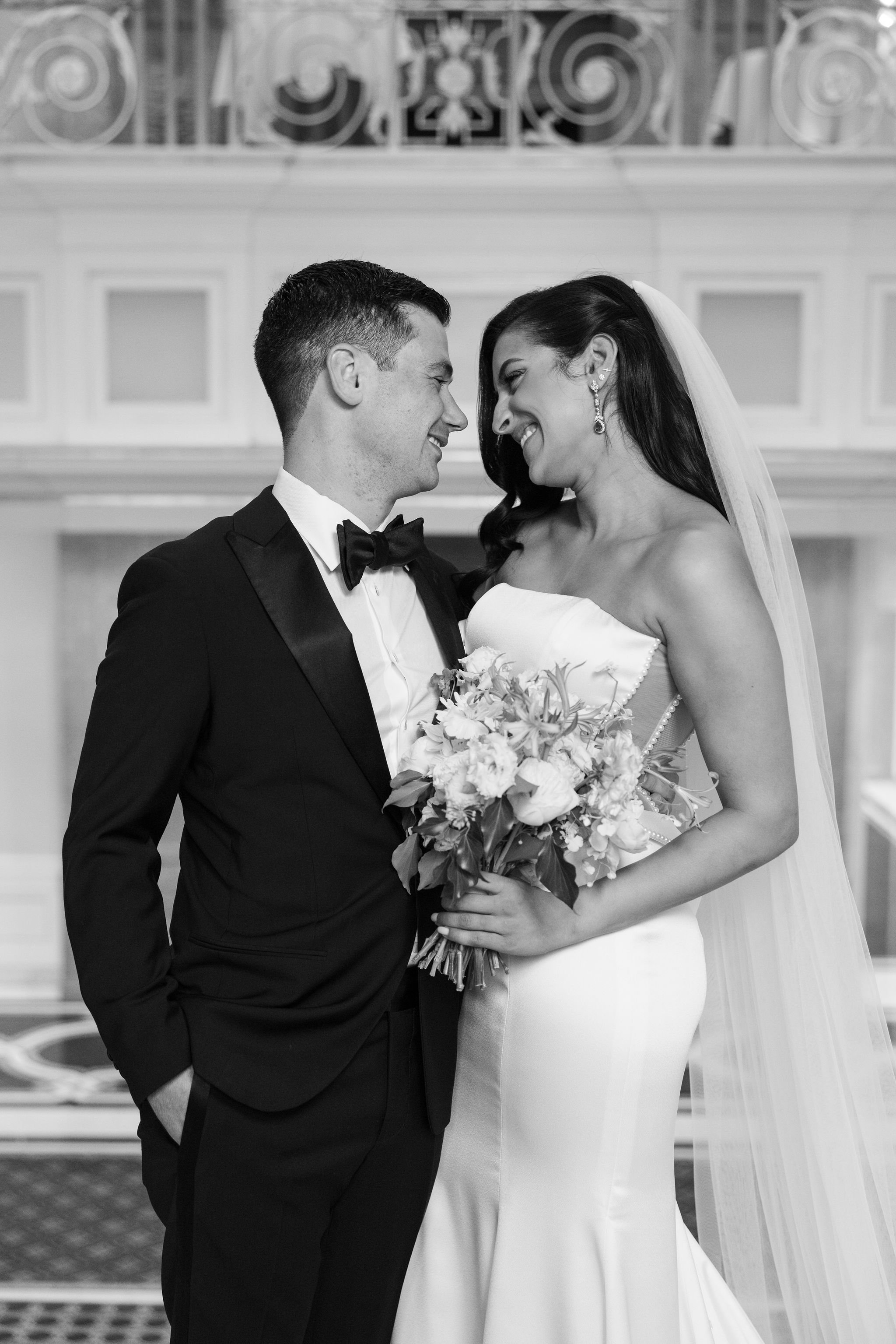 A black and white photo of a bride and groom on their wedding day, facing each other with noses touching, smiling, and holding a bouquet of flowers.
