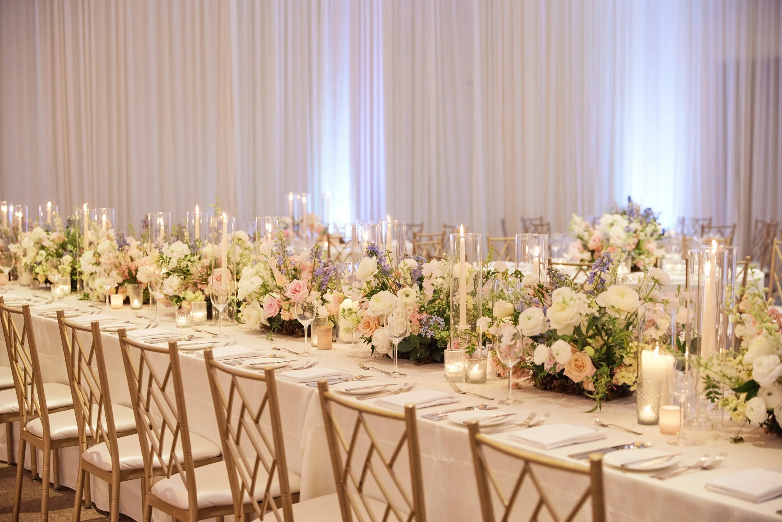Elegant wedding reception setup with a long table decorated with a lush floral centerpiece and candles, chairs with white cushions, and a backdrop of soft, flowing drapes.