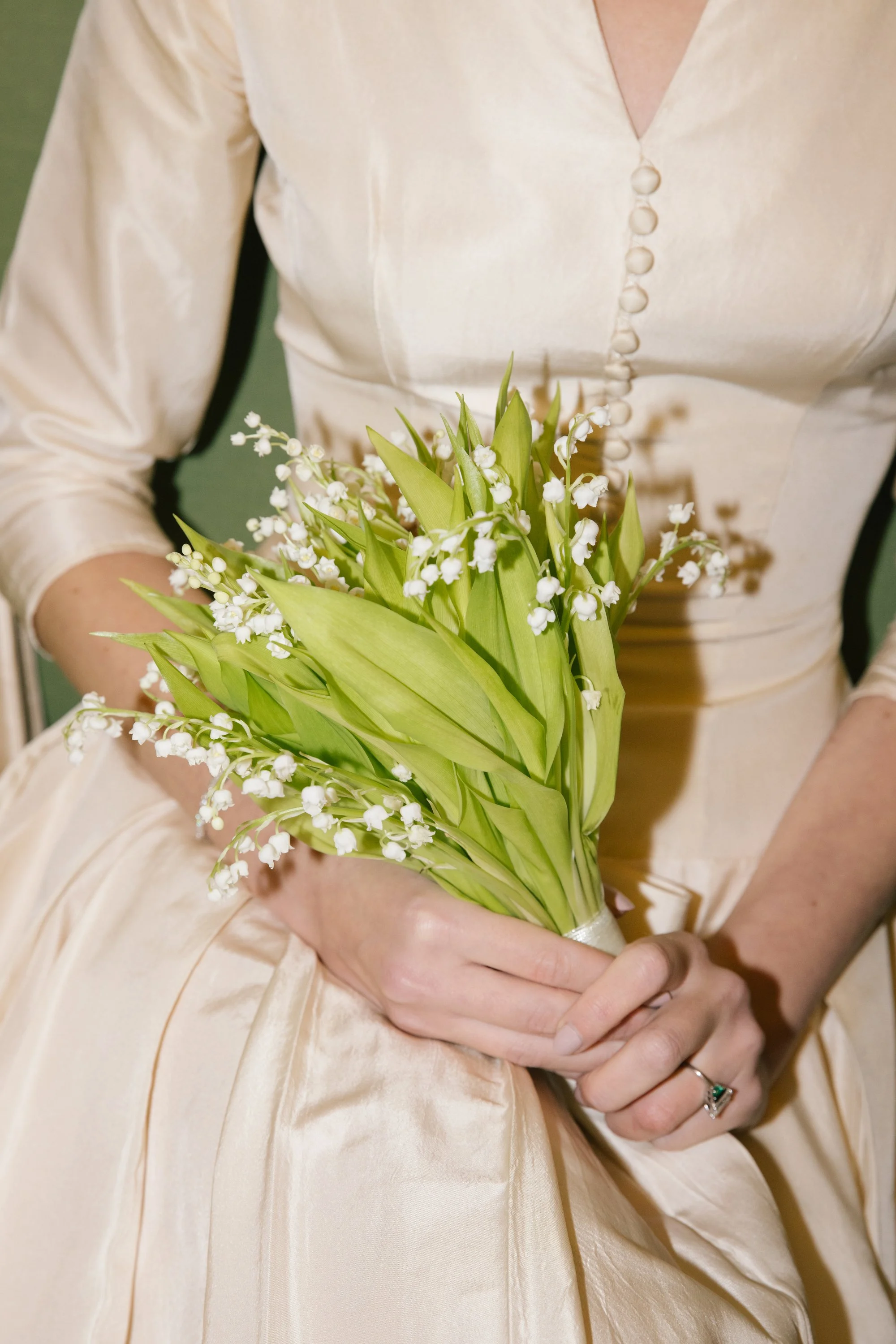 A woman in a cream-colored satin dress is holding a bouquet of white lilies of the valley flowers.