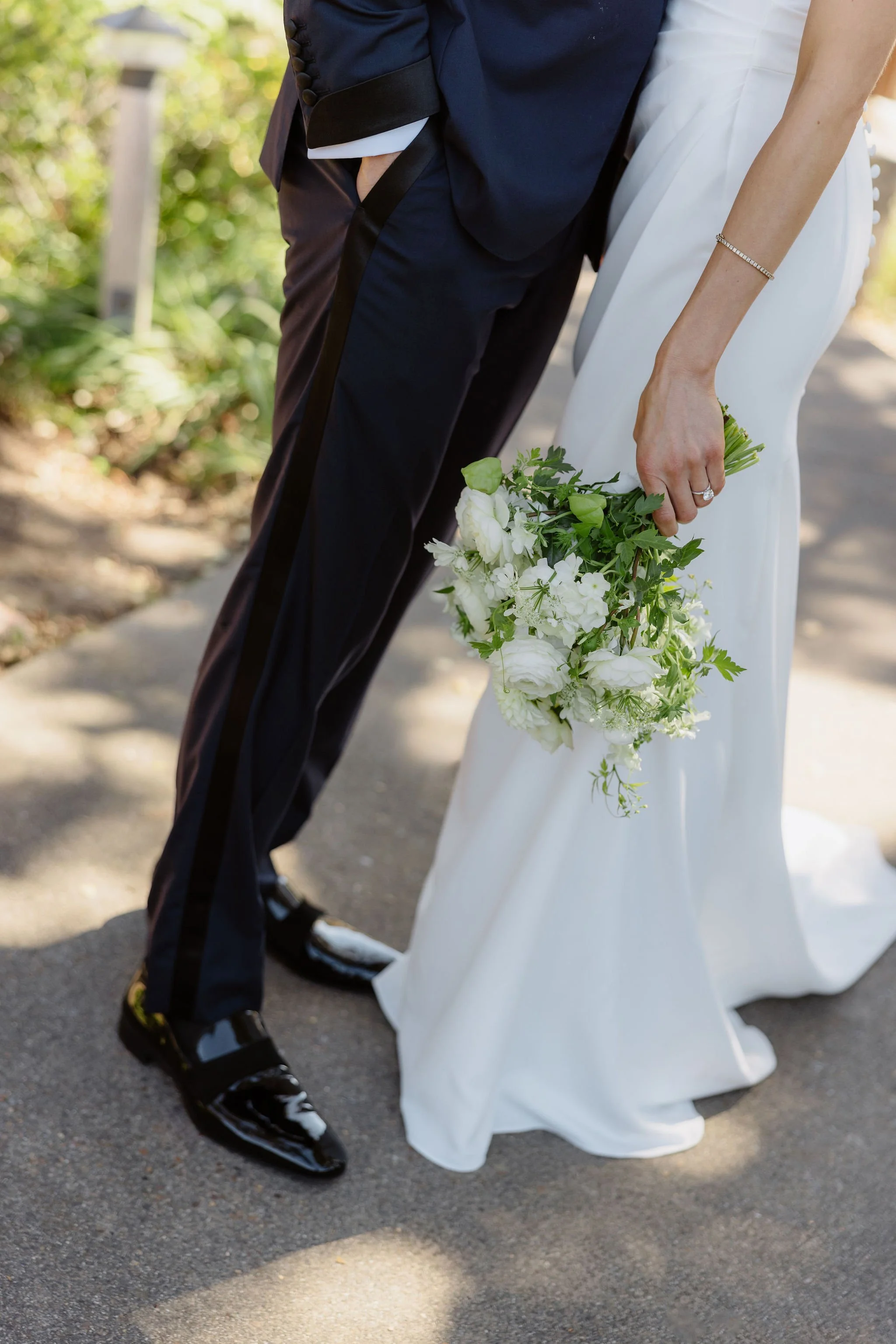 A close-up of a bride and groom holding hands with their bodies slightly turned, the bride in a white wedding gown holding a bouquet of white flowers with greenery, and the groom in a dark navy tuxedo with shiny black shoes, outdoors on a paved path 