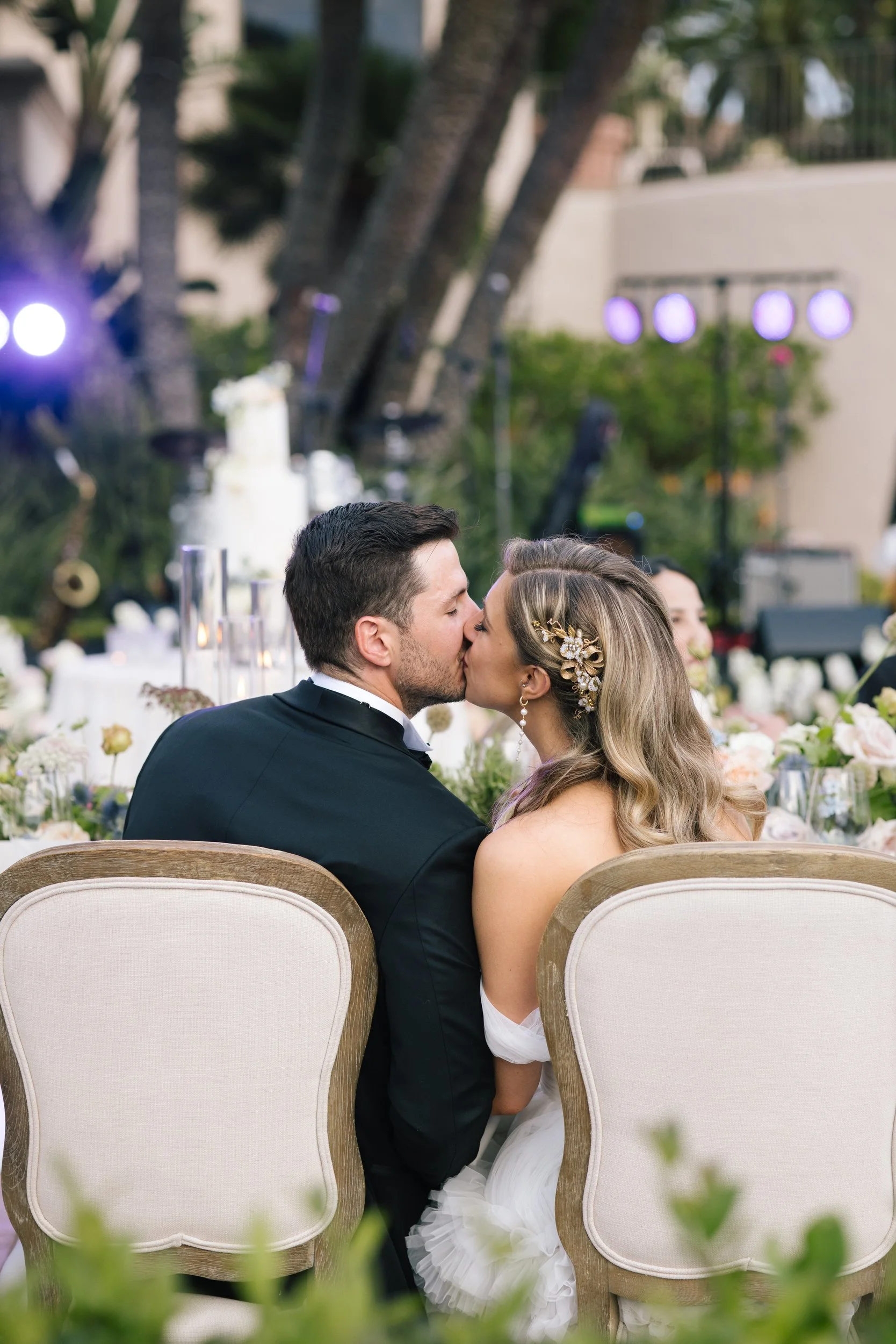 A bride and groom share a kiss during their wedding ceremony outdoors, surrounded by tables with floral arrangements and trees, with union lights and a white building in the background.