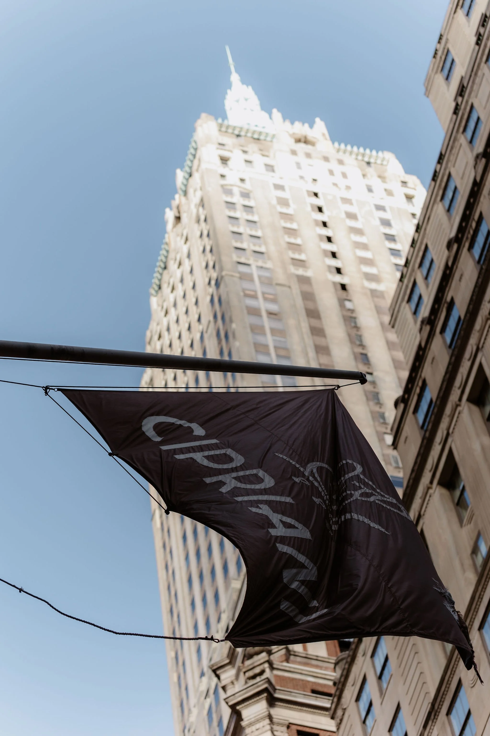 Upward view of a tall skyscraper with a pointed spire, a flagpole, and neighboring buildings under a clear blue sky, with a black flag displaying white text in the foreground.