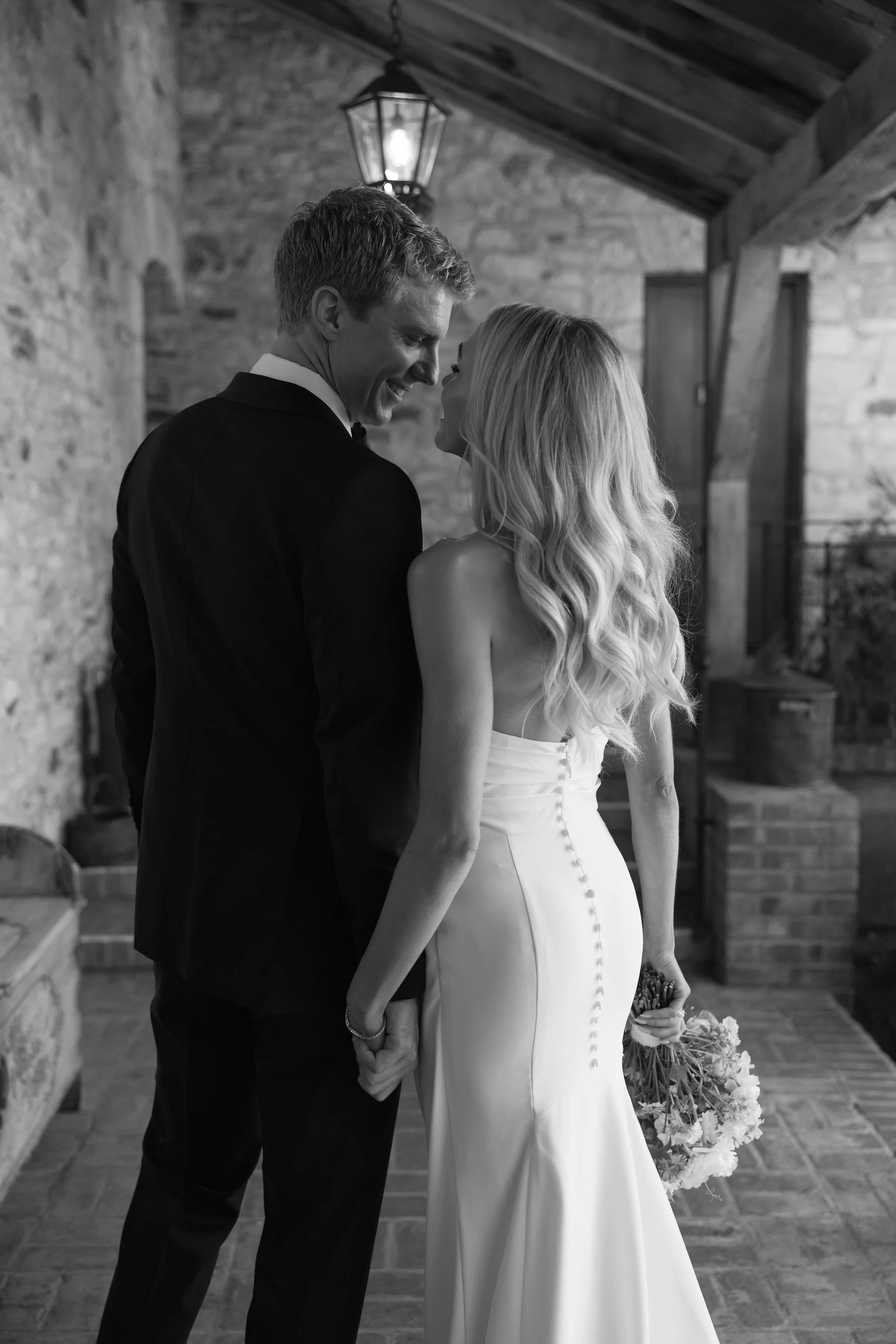 A black and white photo of a bride and groom holding hands and looking at each other in a rustic indoor setting, with stone walls, wooden beams, and a hanging lantern ceiling light.