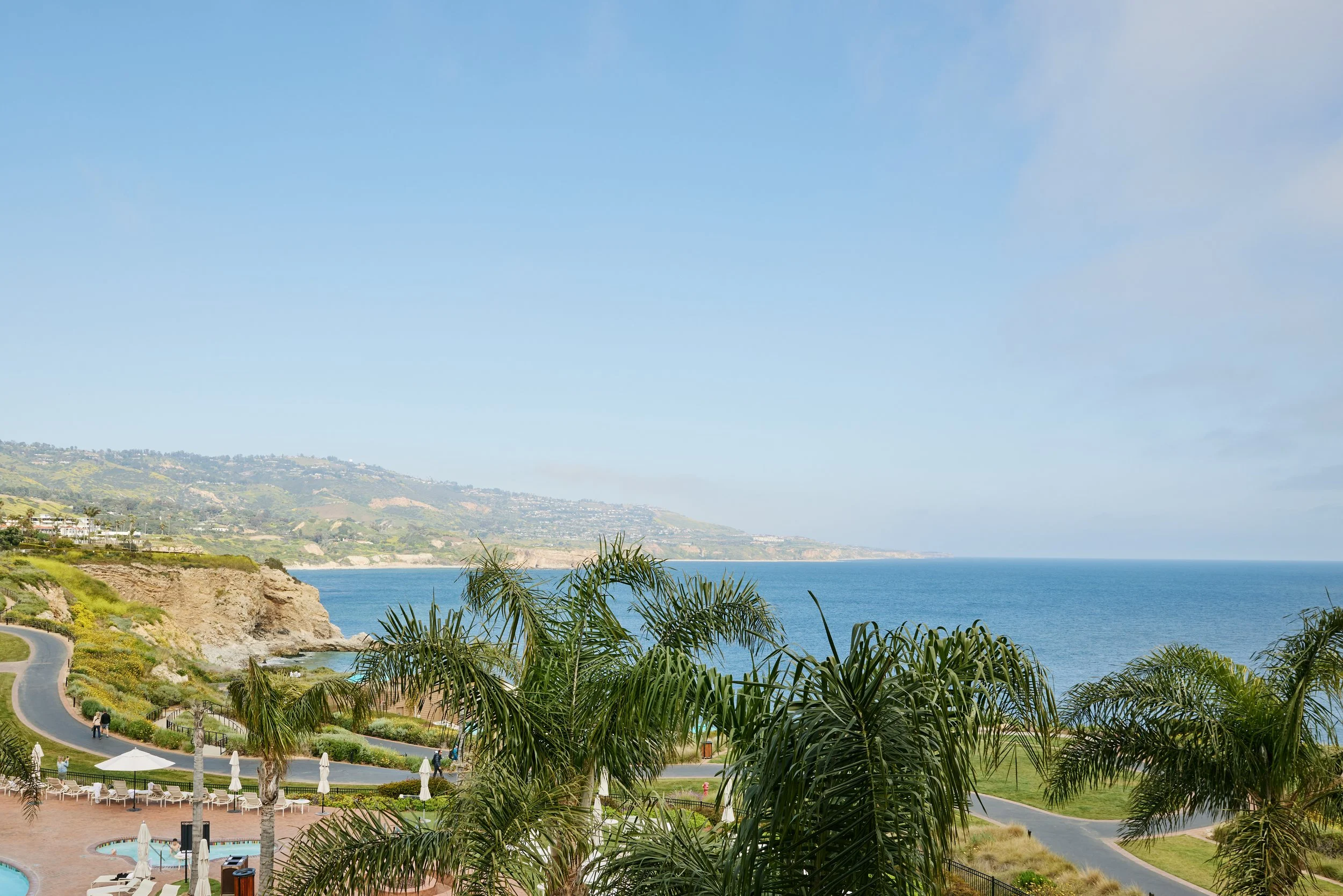 View of a coastal landscape with cliffs, the ocean, and a park area with palm trees and outdoor seating.