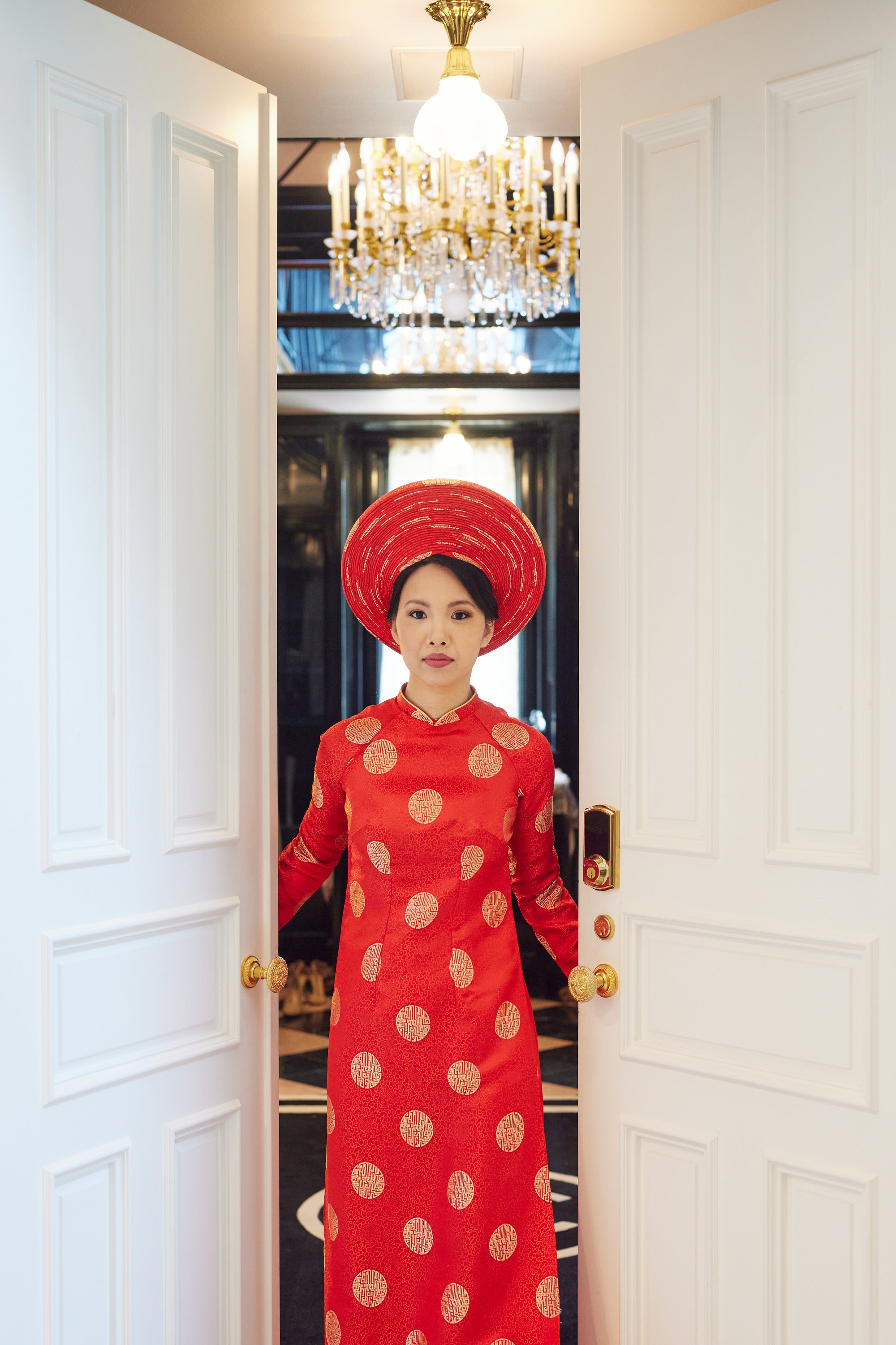 A woman dressed in traditional red Vietnamese áo dài with gold circular patterns, wearing a large red conical hat, standing in a doorway with open white doors, in a room decorated with a large chandelier.