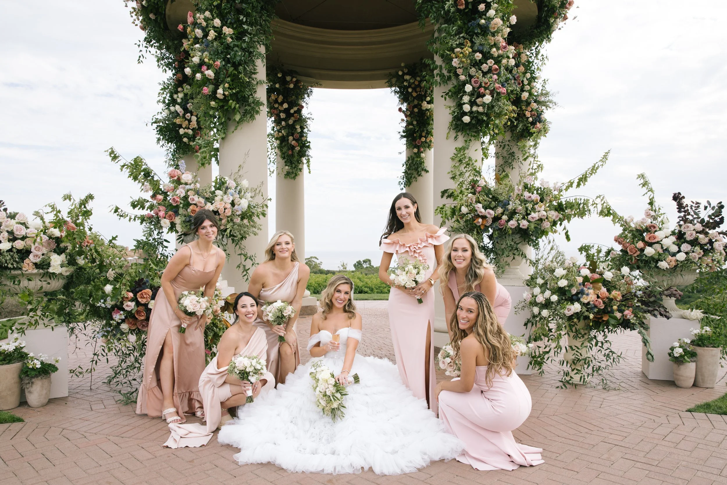 A group of women, likely bridesmaids, posing for a photo at a wedding outdoors, with a floral decorated structure in the background.