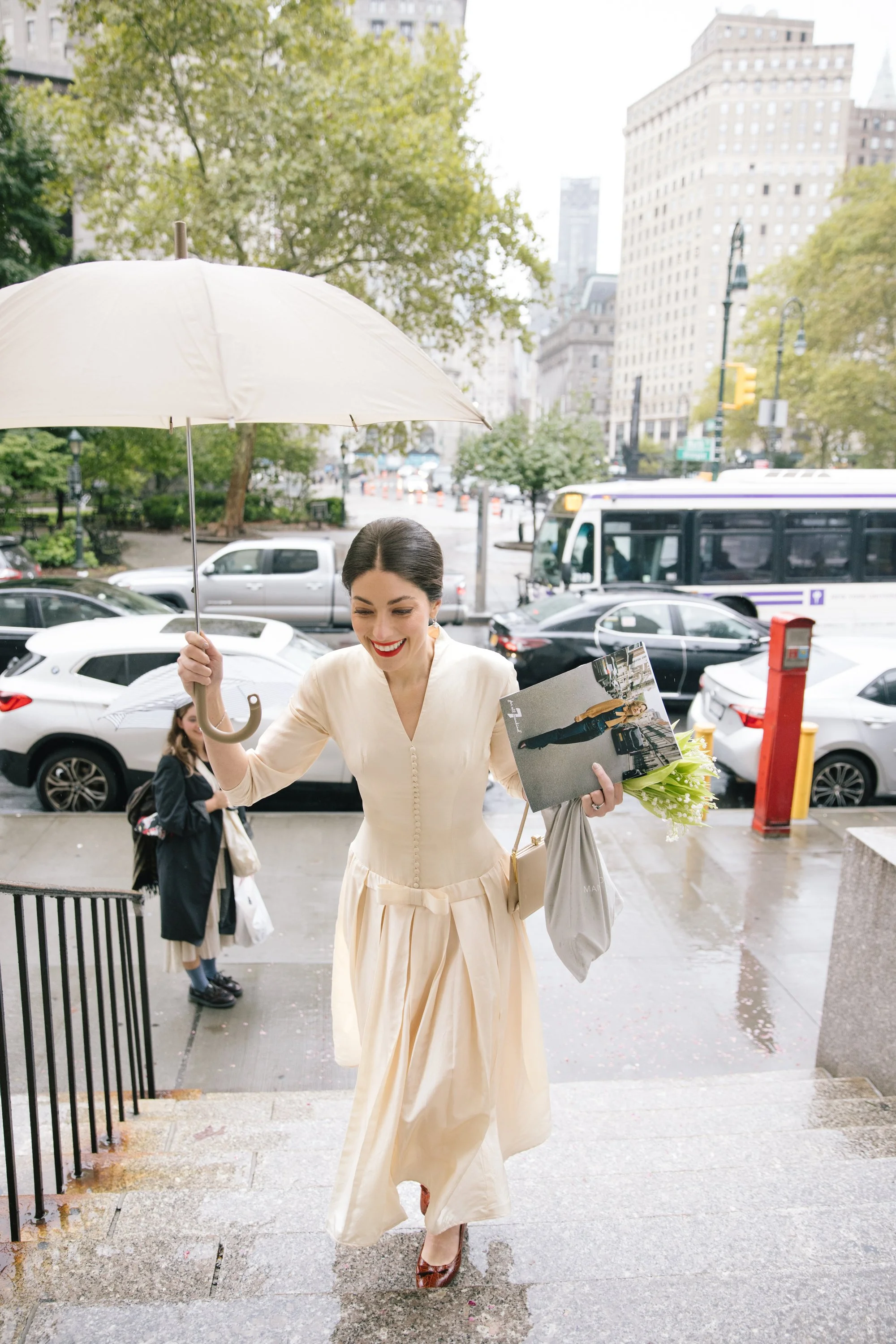 A woman in a cream-colored dress holding a bouquet of flowers and a magazine, smiling while walking out of a building on a rainy day, with city buildings, trees, and parked cars in the background.