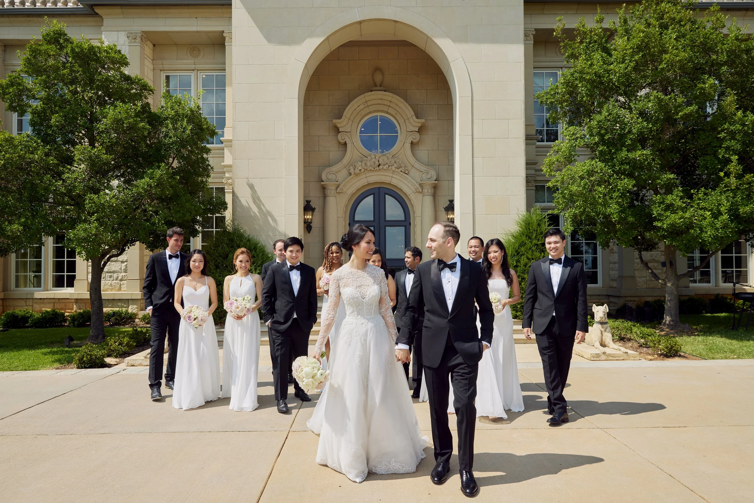 A bride and groom walk hand in hand outside a building with architectural details, surrounded by wedding party members dressed in formal attire, with greenery and trees in the background.