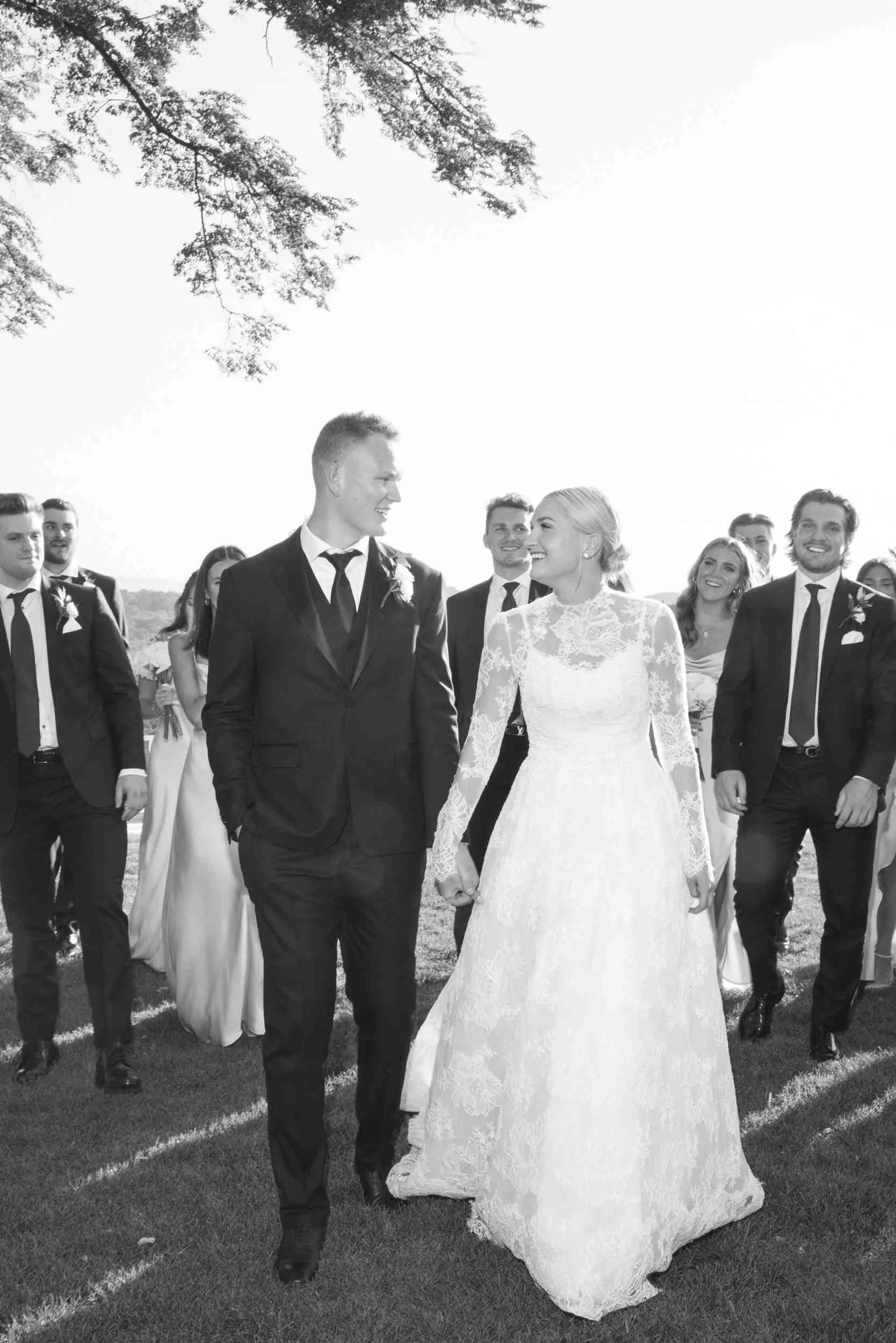 Black and white photo of a bride and groom holding hands and walking outdoors, surrounded by smiling friends in formal attire.