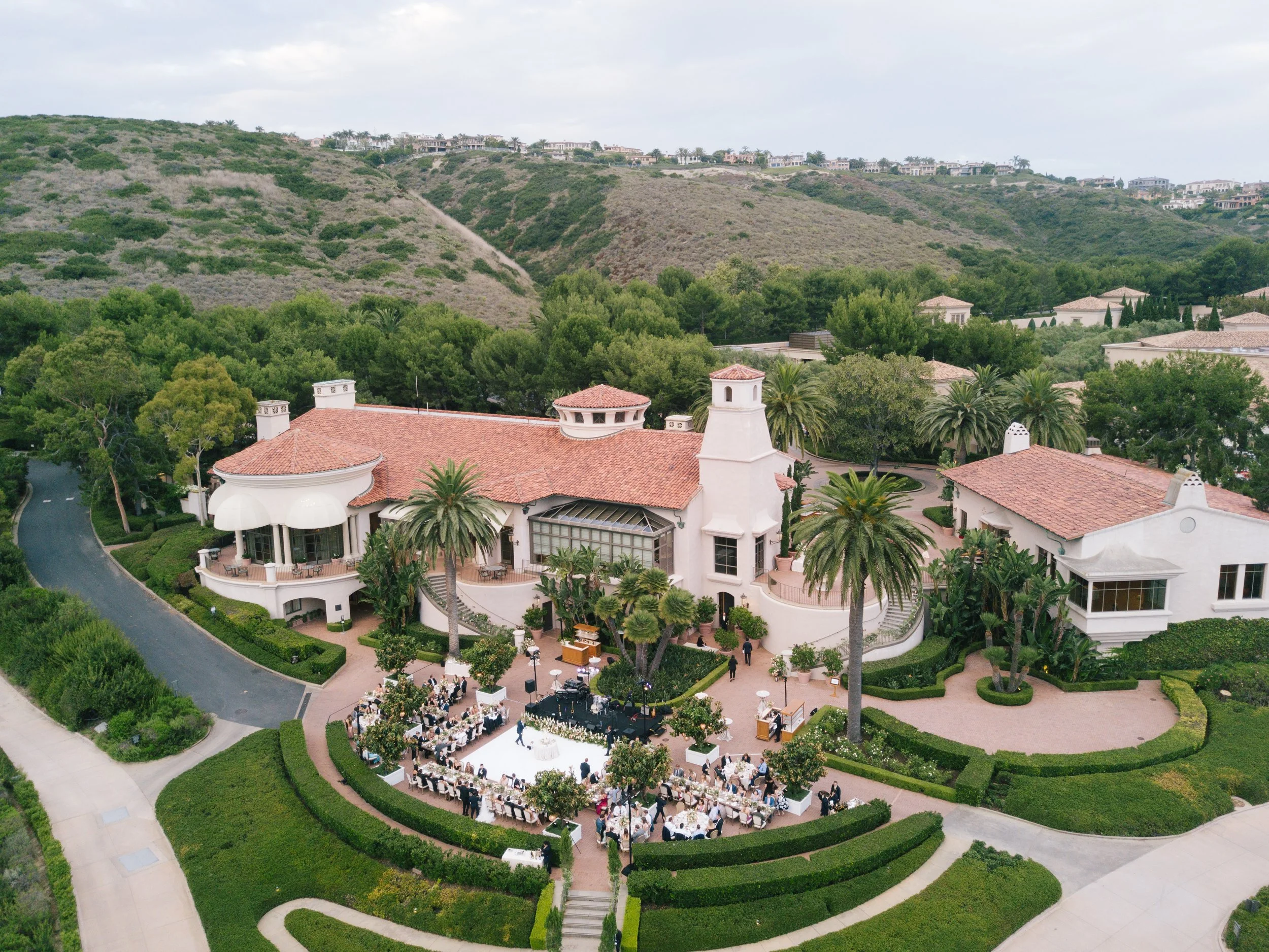 A large outdoor wedding ceremony with guests seated in front of a white aisle, set within a lush garden surrounded by manicured hedges and tall trees. The ceremony is taking place on a circular platform under an open sky, at the center of a courtyard