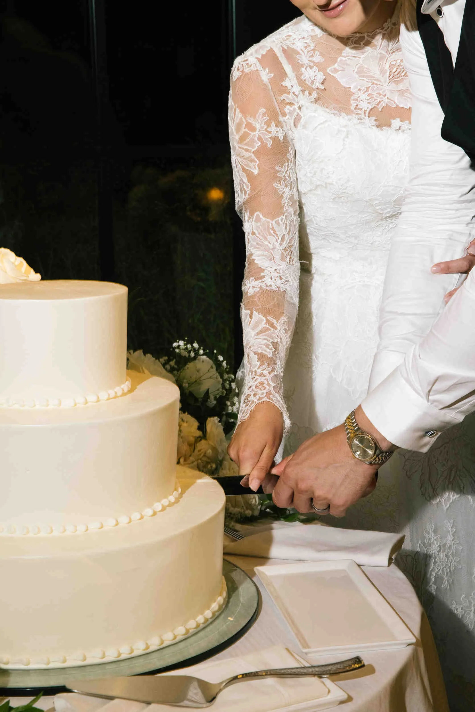 A couple cutting a wedding cake at their wedding reception. The bride is wearing a lace wedding dress, and the groom is dressed in a white shirt and black vest. The three-tiered wedding cake is decorated with small white icing pearls.