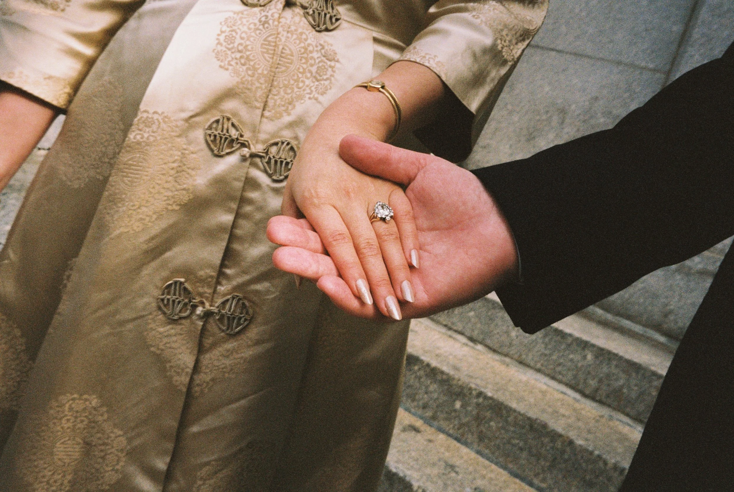 A close-up of two people holding hands, one person wearing a large diamond ring and a gold bracelet, the other person wearing a ring and a black sleeve, with steps in the background.