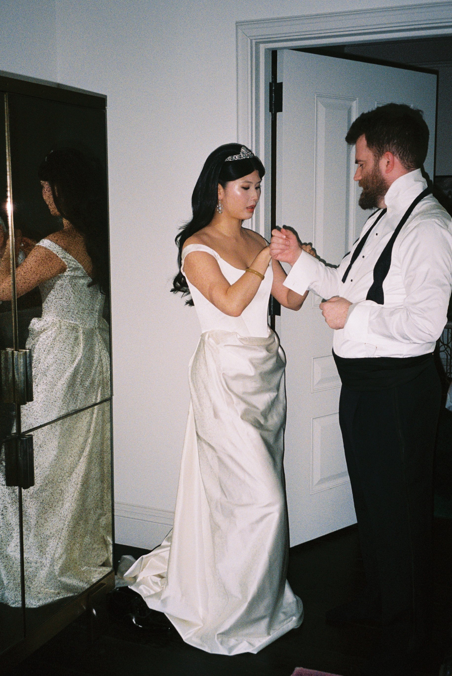 A bride and groom holding hands in a room, with a mirror reflecting the bride's dress, a door in the background, and a dark floor.