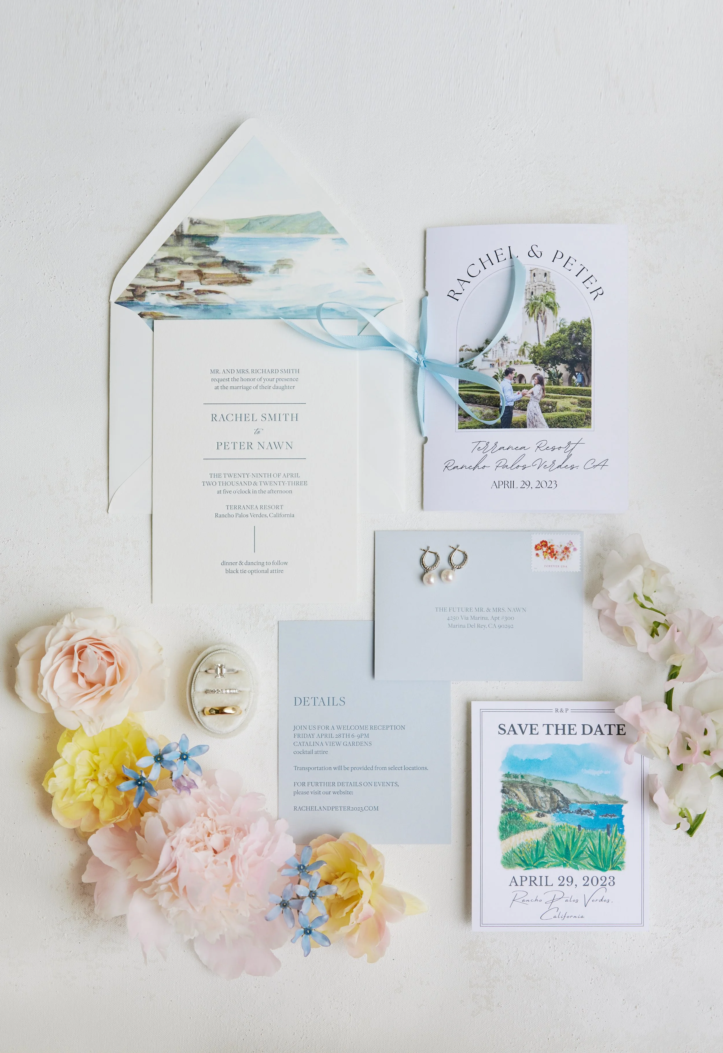 Wedding invitation suite with floral decorations, including circular and rectangular cards, a save the date, and jewelry, all arranged on a white surface with white, pink, yellow, and blue flowers.