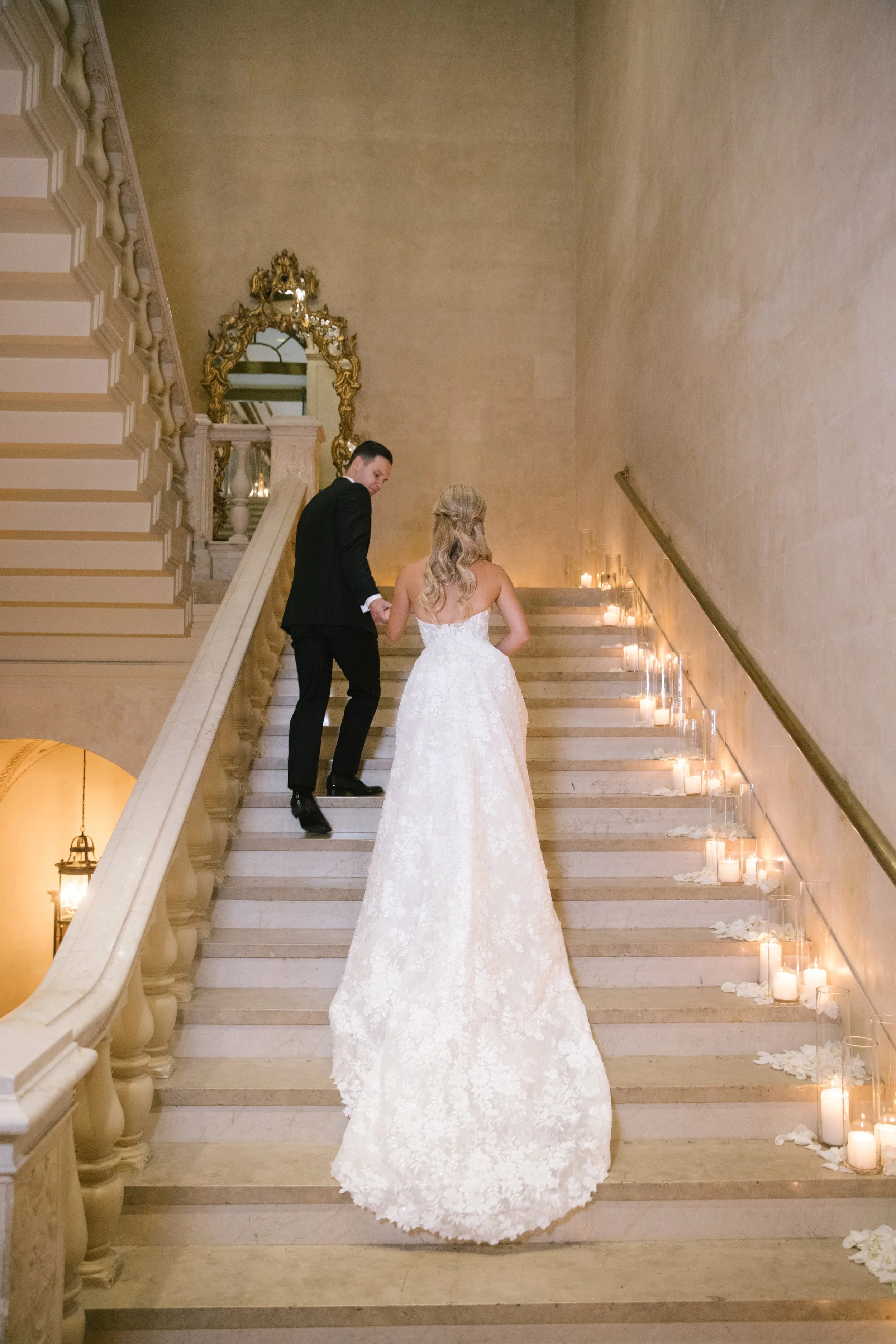 A bride and groom holding hands while walking up a staircase decorated with candles and flower petals, with ornate mirrors and gold accents in the background.