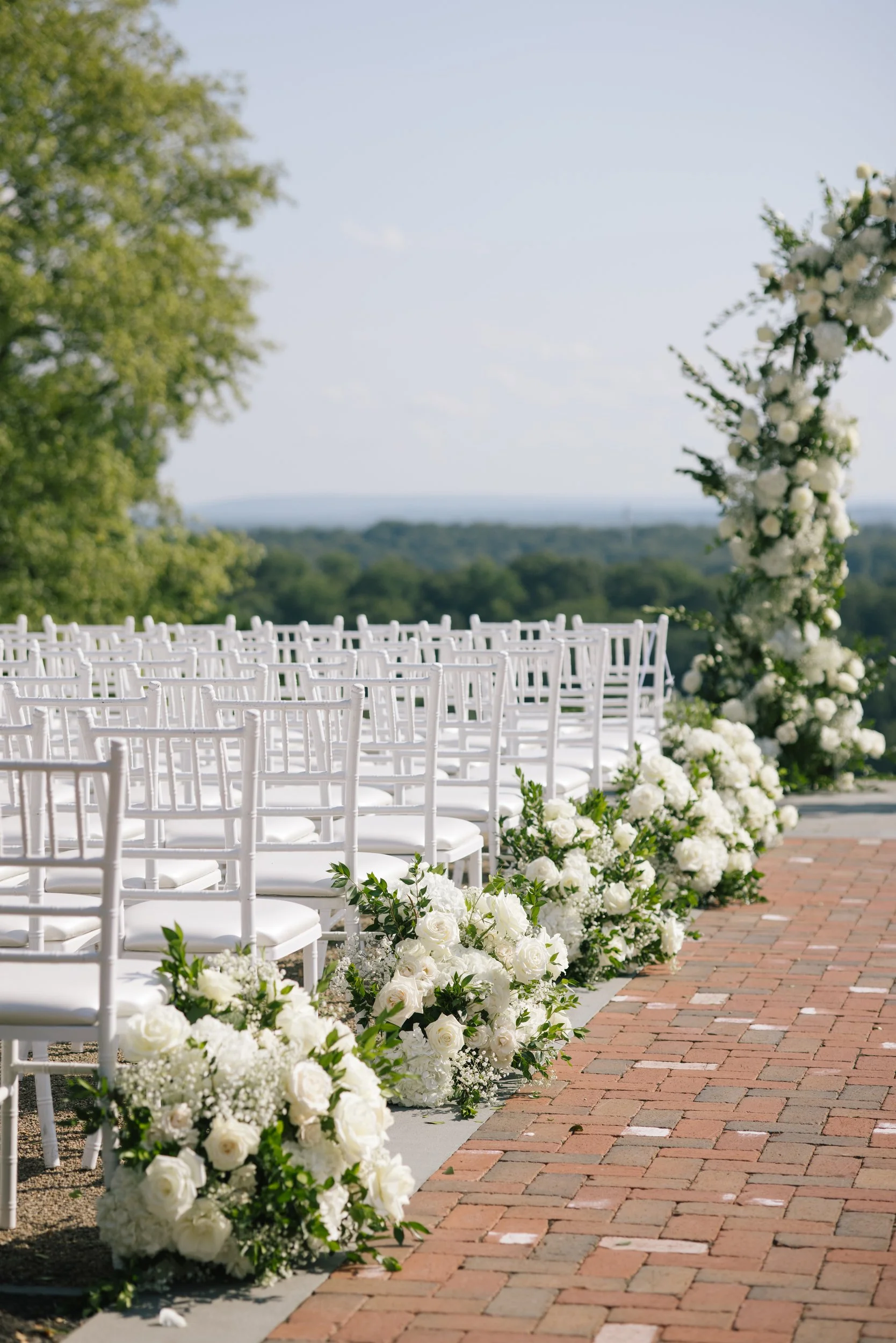 Outdoor wedding ceremony setup with white chairs and floral arrangements along a brick pathway, under a tree with a scenic landscape in the background.