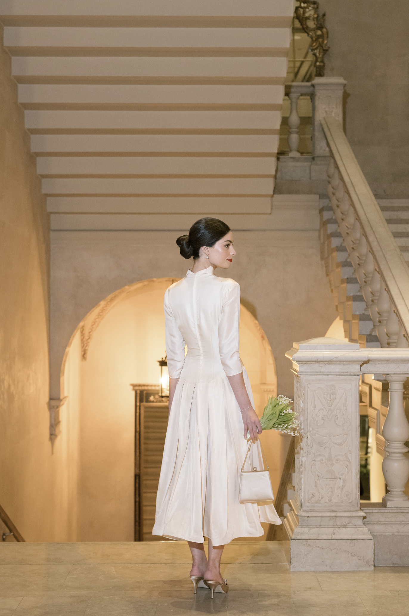 A woman dressed in a white vintage-style gown holding a bouquet of white flowers, standing on a staircase in an elegant, classic interior with warm lighting.