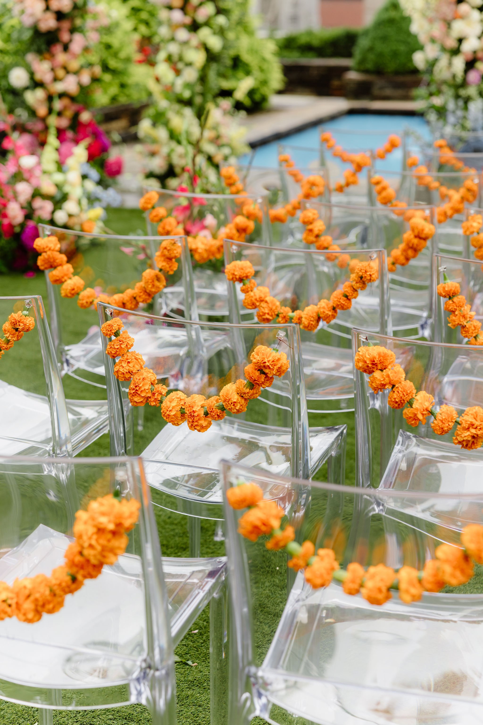 Clear chairs decorated with orange flower garlands, arranged outdoors on grass, surrounding a white table with floral arrangements in the background.