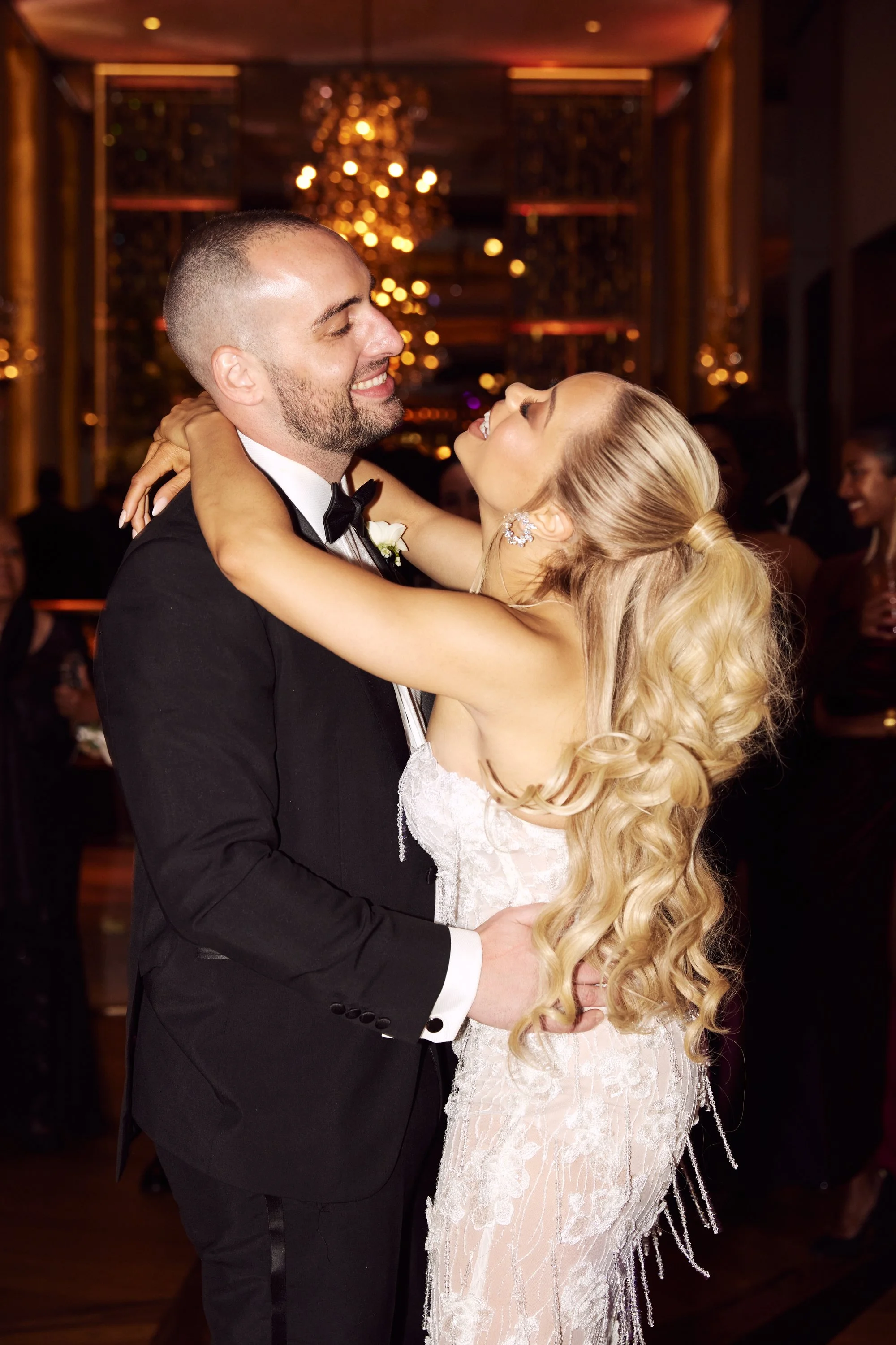A man and a woman are dancing closely at a formal event, with the man wearing a tuxedo and the woman in a white lace dress. They are smiling and looking into each other’s eyes, surrounded by other guests in the background and warm, elegant lighting.