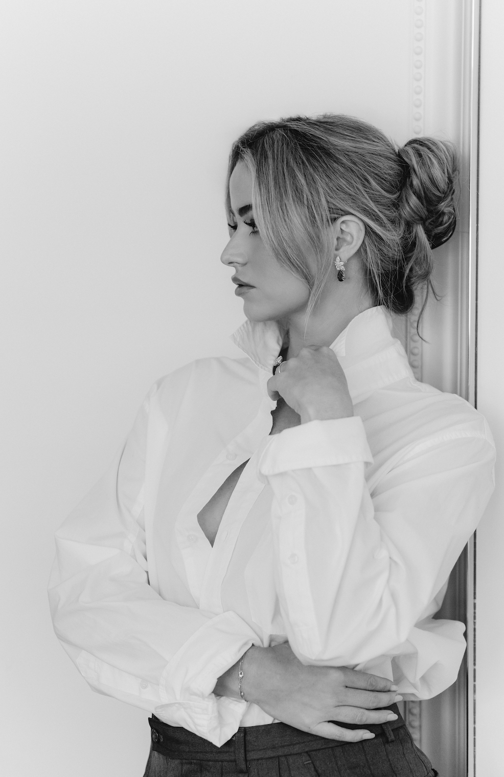 A black-and-white photo of a woman with styled hair, wearing earrings and a white blouse, standing against a plain wall with her hand on her collar.