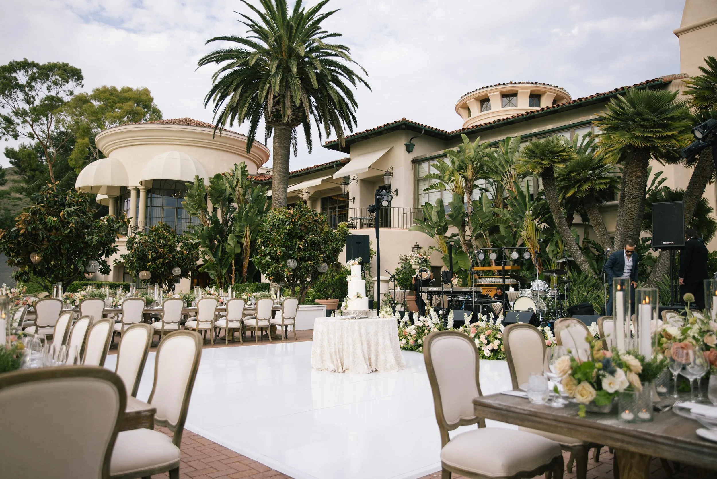 Outdoor wedding setup with decorated tables, floral arrangements, and a cake in front of a large house with palm trees and a scenic sky.