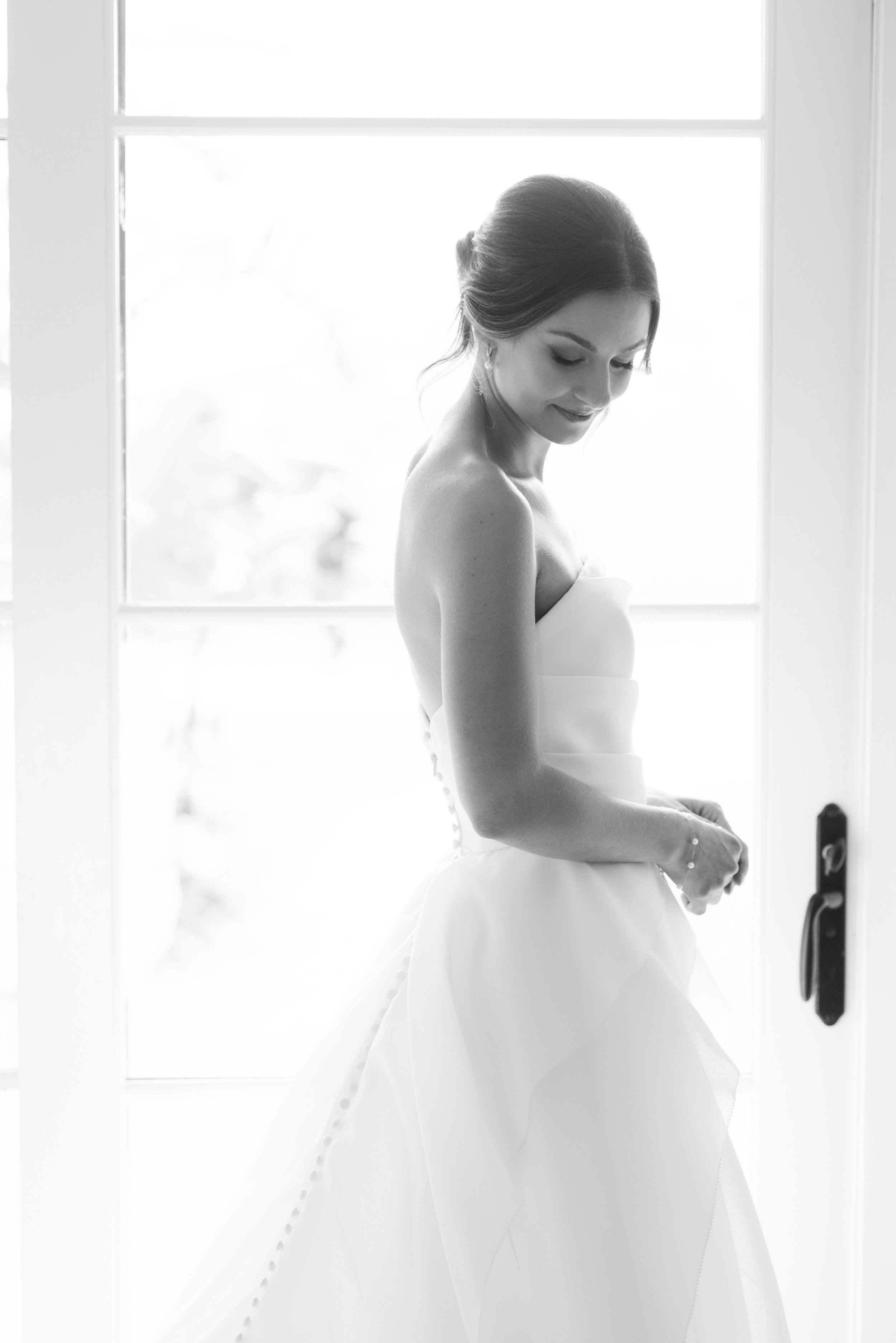 Black and white photo of a woman in a wedding dress standing by a white door with a window, her head tilted down slightly, with natural light coming from the window behind her.