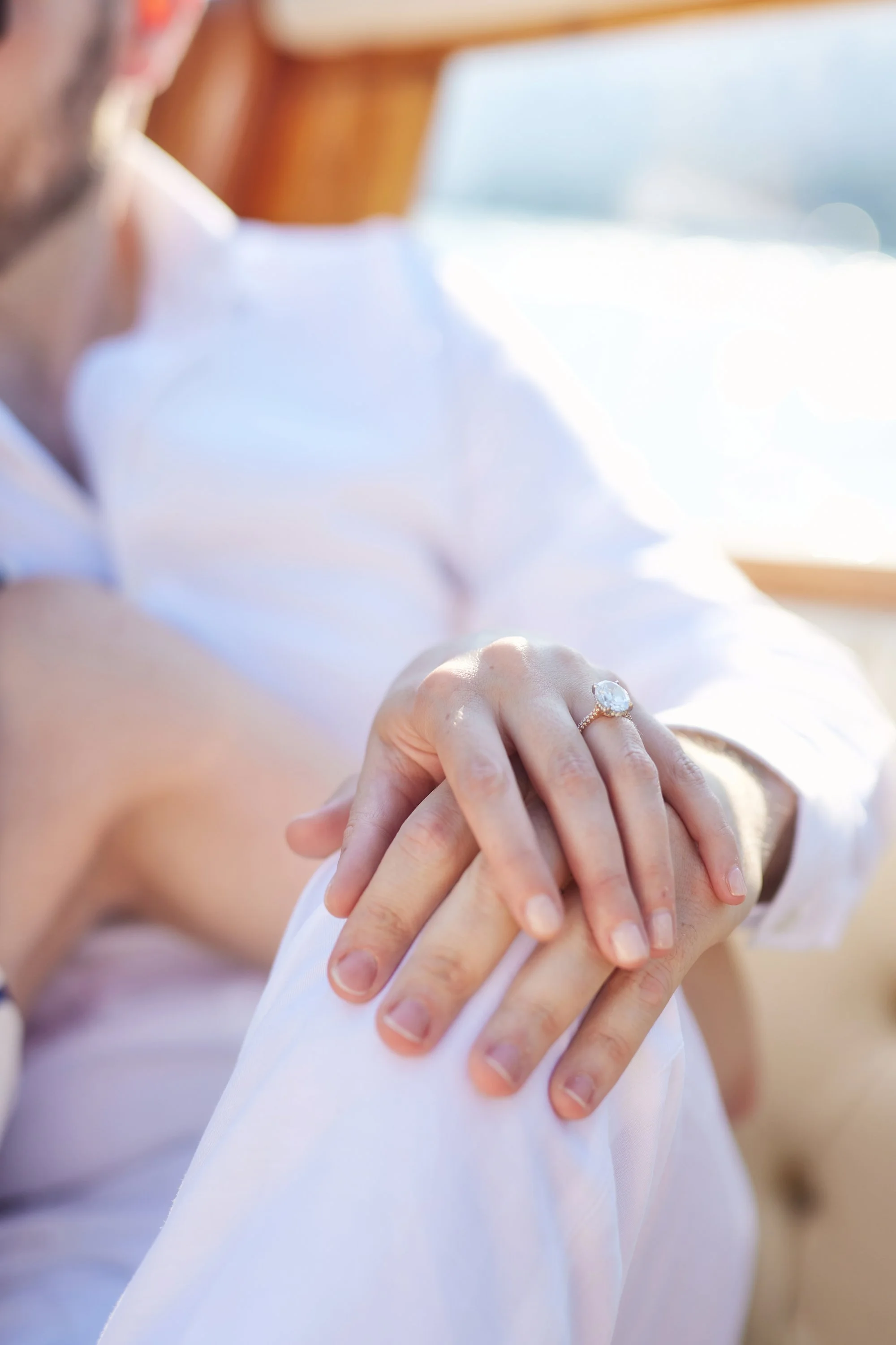 A woman wearing a wedding ring looks at her hand with her fingers delicately touching her arm, near a window with bright natural light, suggesting an intimate or joyous moment.