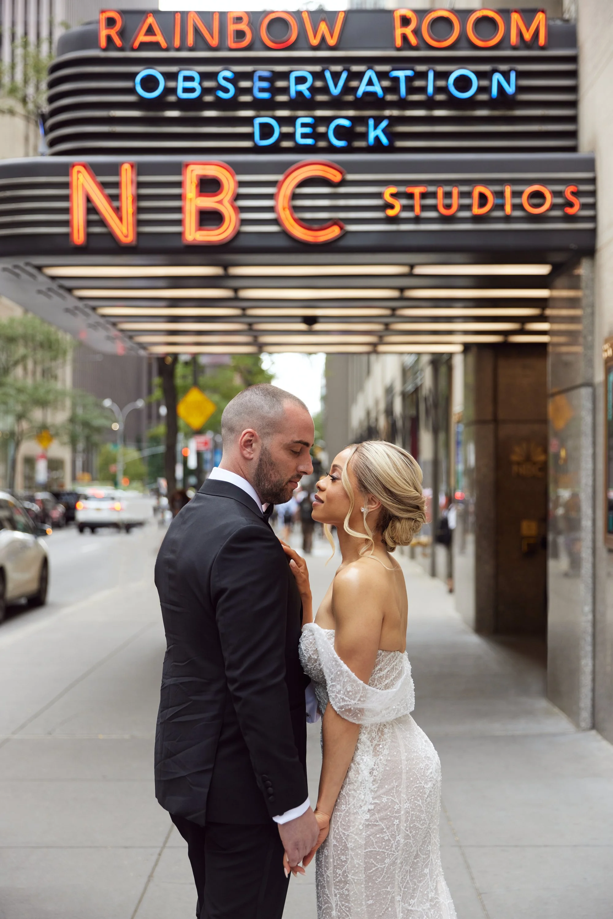 A bride and groom stand closely together on a city sidewalk, looking into each other's eyes. The bride wears a white lace wedding gown with an off-shoulder design, and the groom is dressed in a black suit. Behind them is a building with a neon sign r