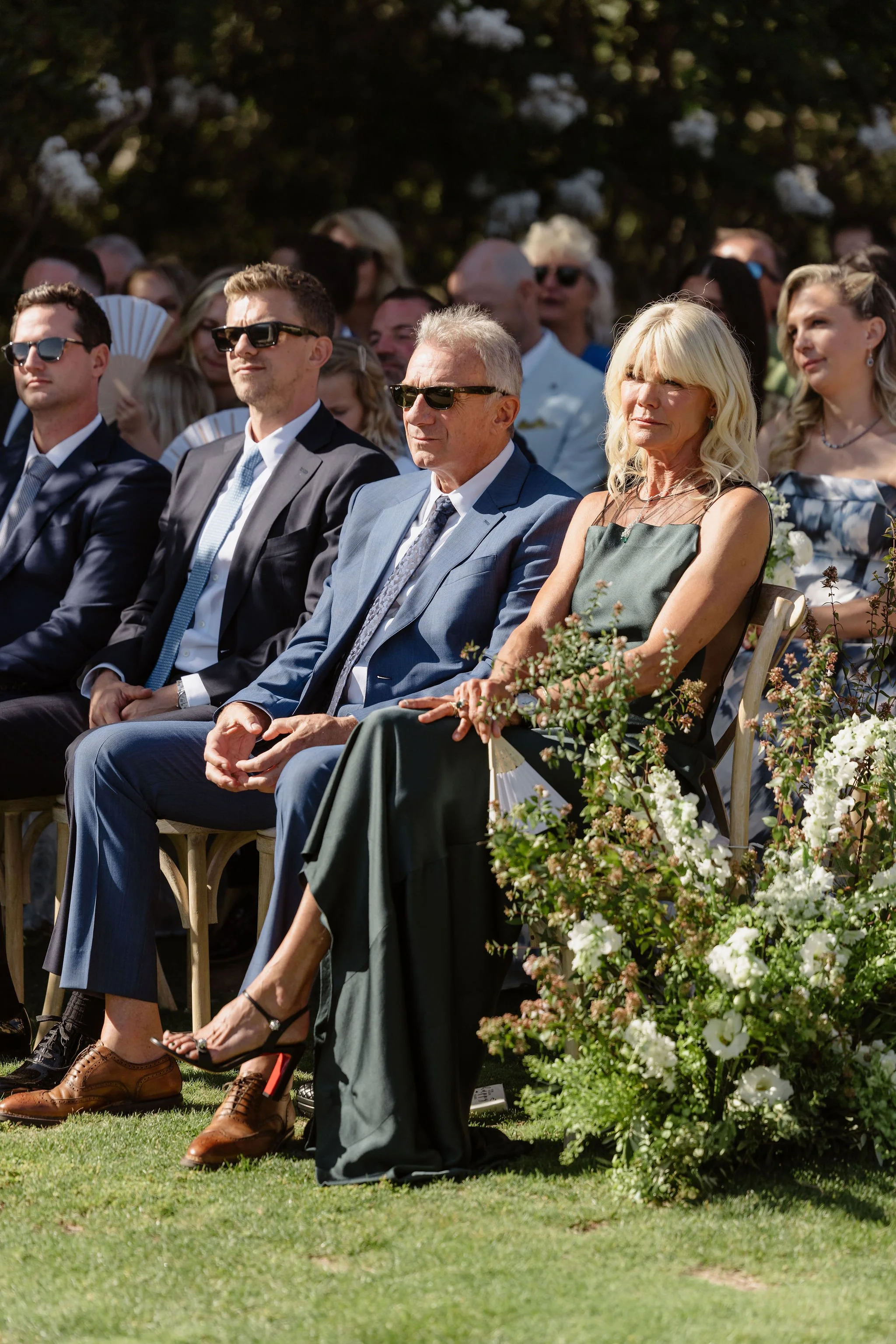 A group of people sitting outdoors in formal attire, attending a wedding or similar event, with floral decorations in the foreground and trees in the background.