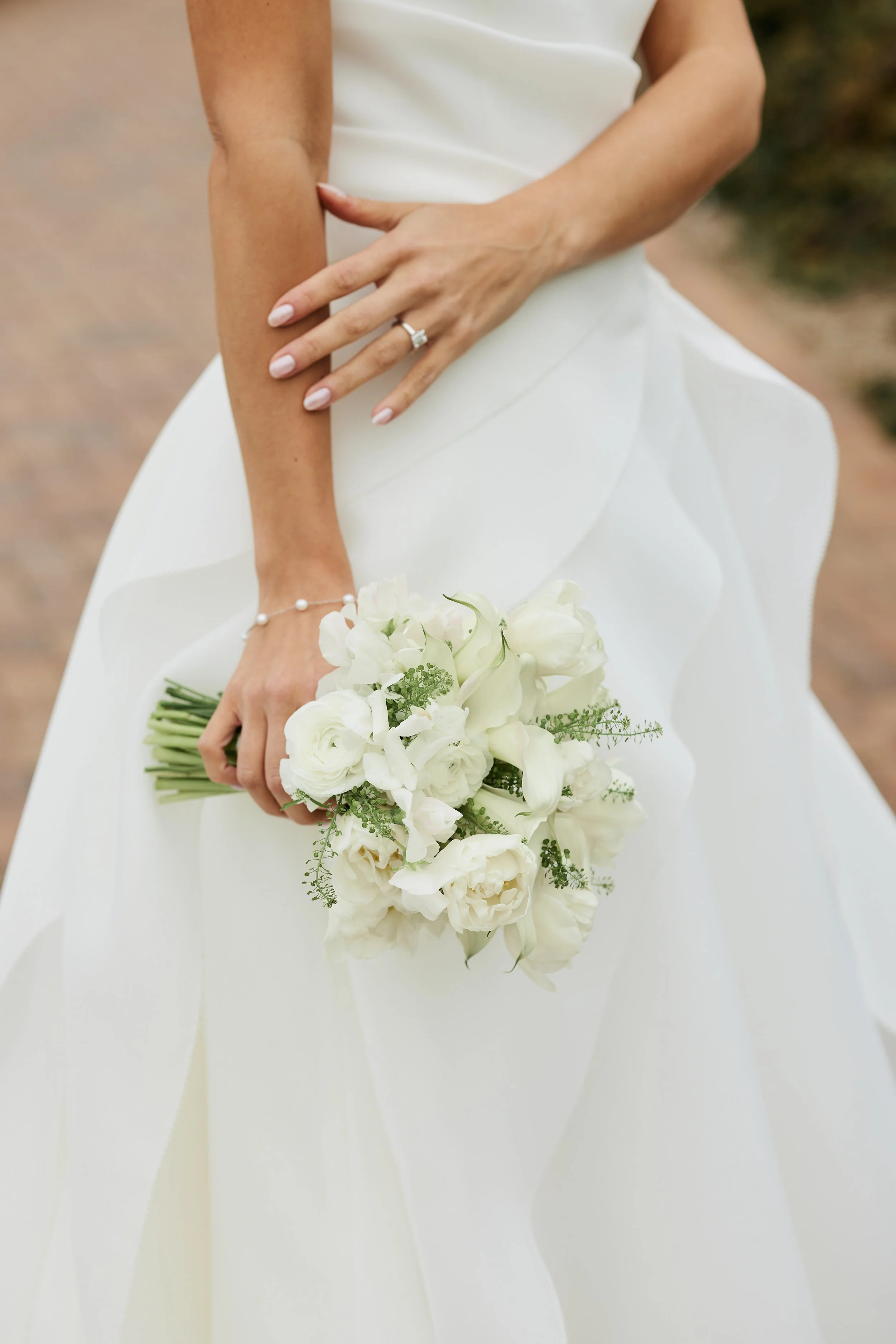 Close-up of a woman in a white dress holding a bouquet of white flowers, wearing a diamond ring and a pearl bracelet, outdoors.