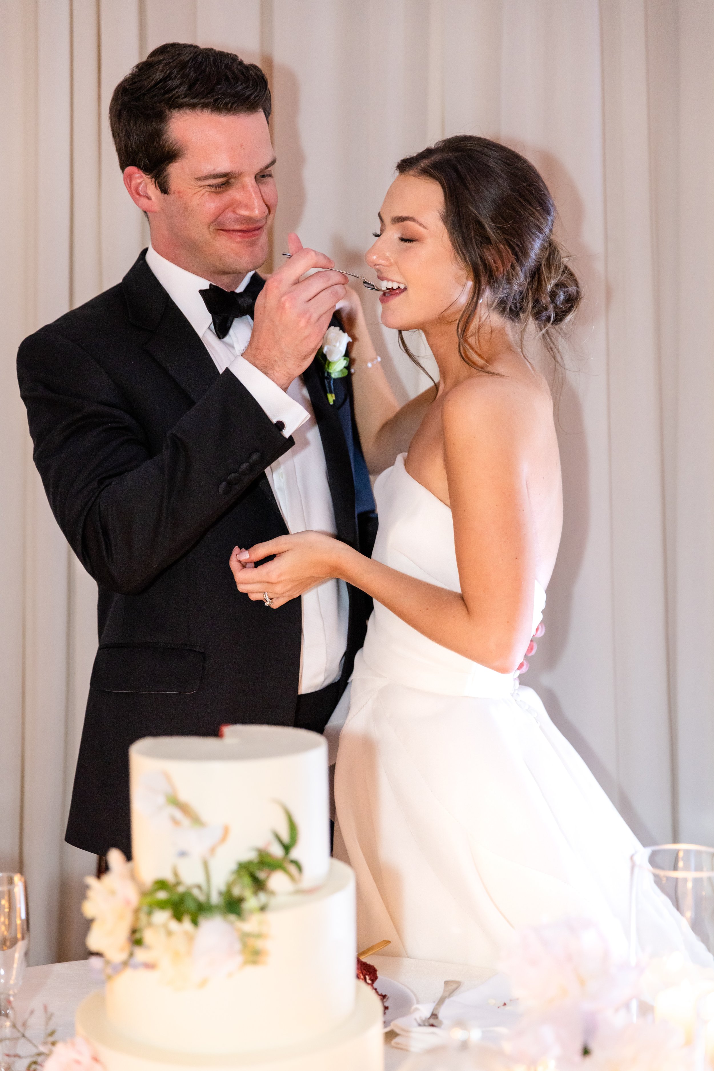 A groom feeding a bride at their wedding reception. The groom is dressed in a tuxedo and the bride is in a strapless wedding gown. They are smiling and holding hands. A wedding cake and floral arrangements are visible in the foreground.