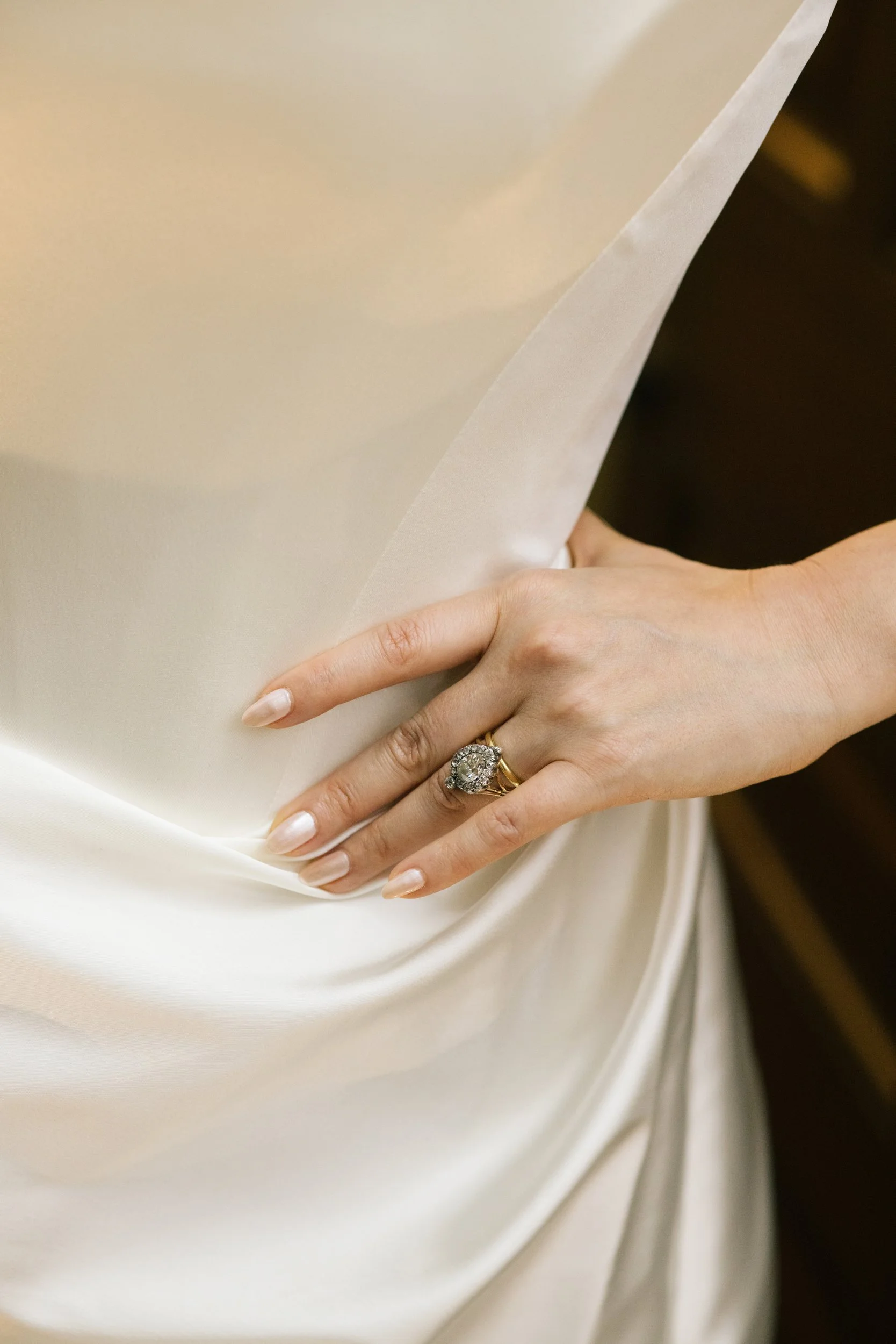 A woman's hand with an ornate ring rests on a cream-colored satin fabric.