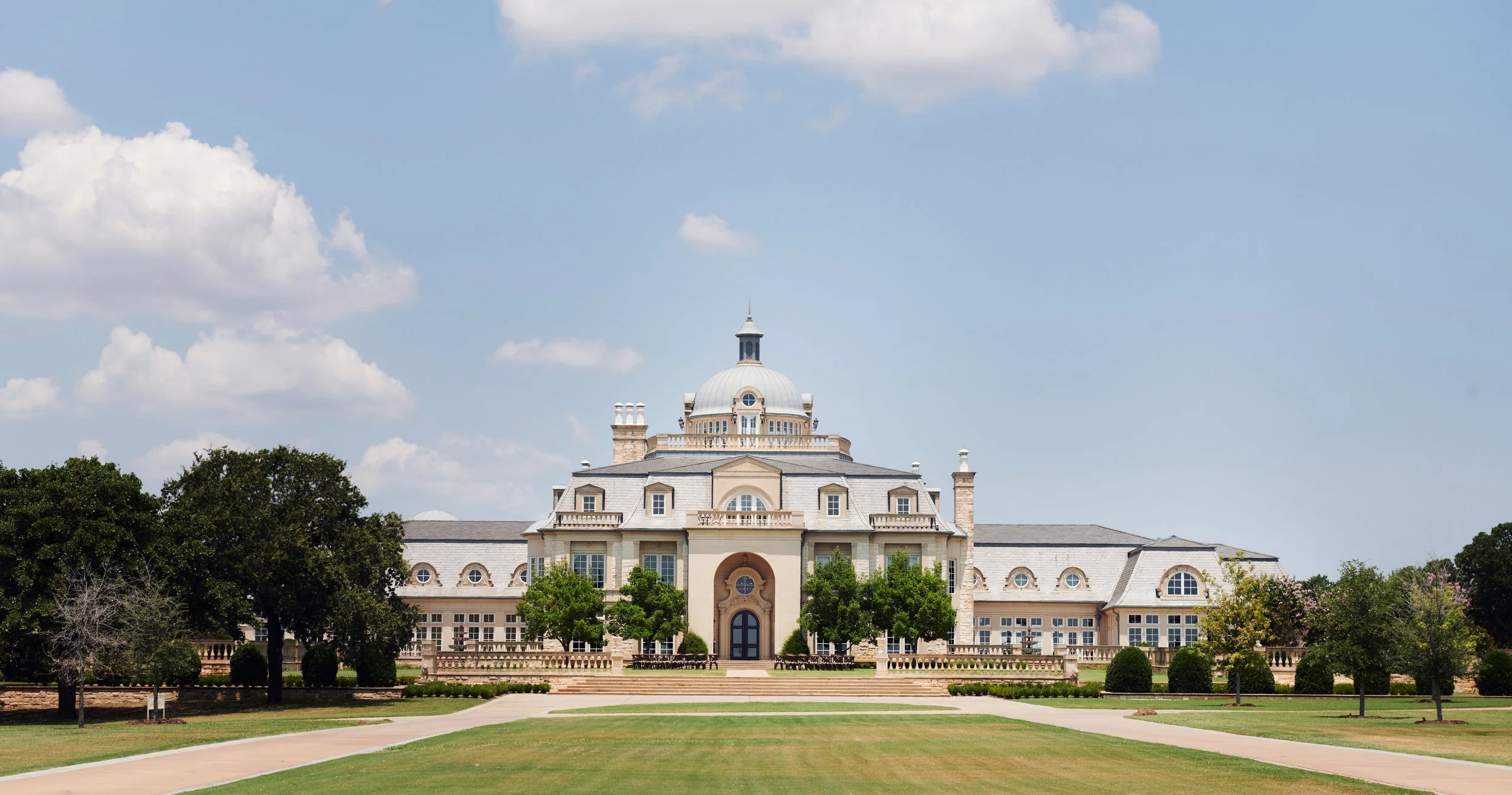 Large, elegant mansion with domed roof, surrounded by trees and a well-maintained lawn under a partly cloudy sky.