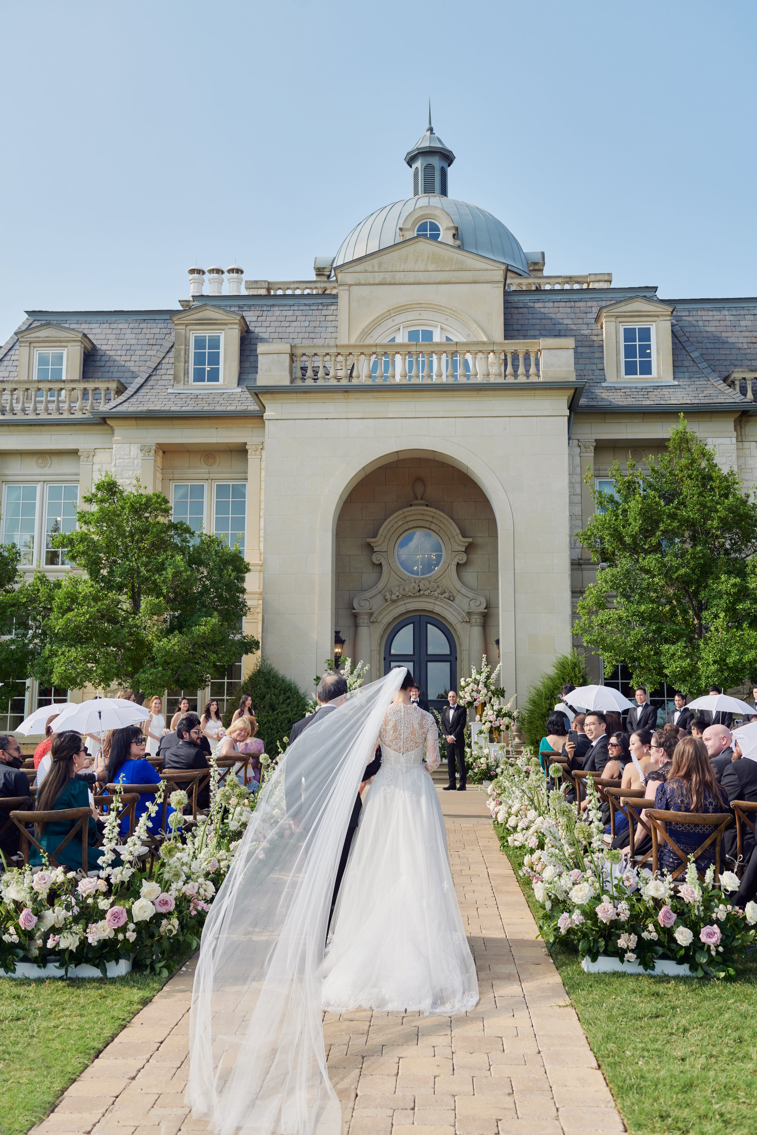Wedding ceremony outdoors in front of a large mansion with a domed roof. The bride and groom walk down the aisle, surrounded by guests seated on both sides, with floral arrangements along the aisle. The bride wears a white dress and veil, and the gue