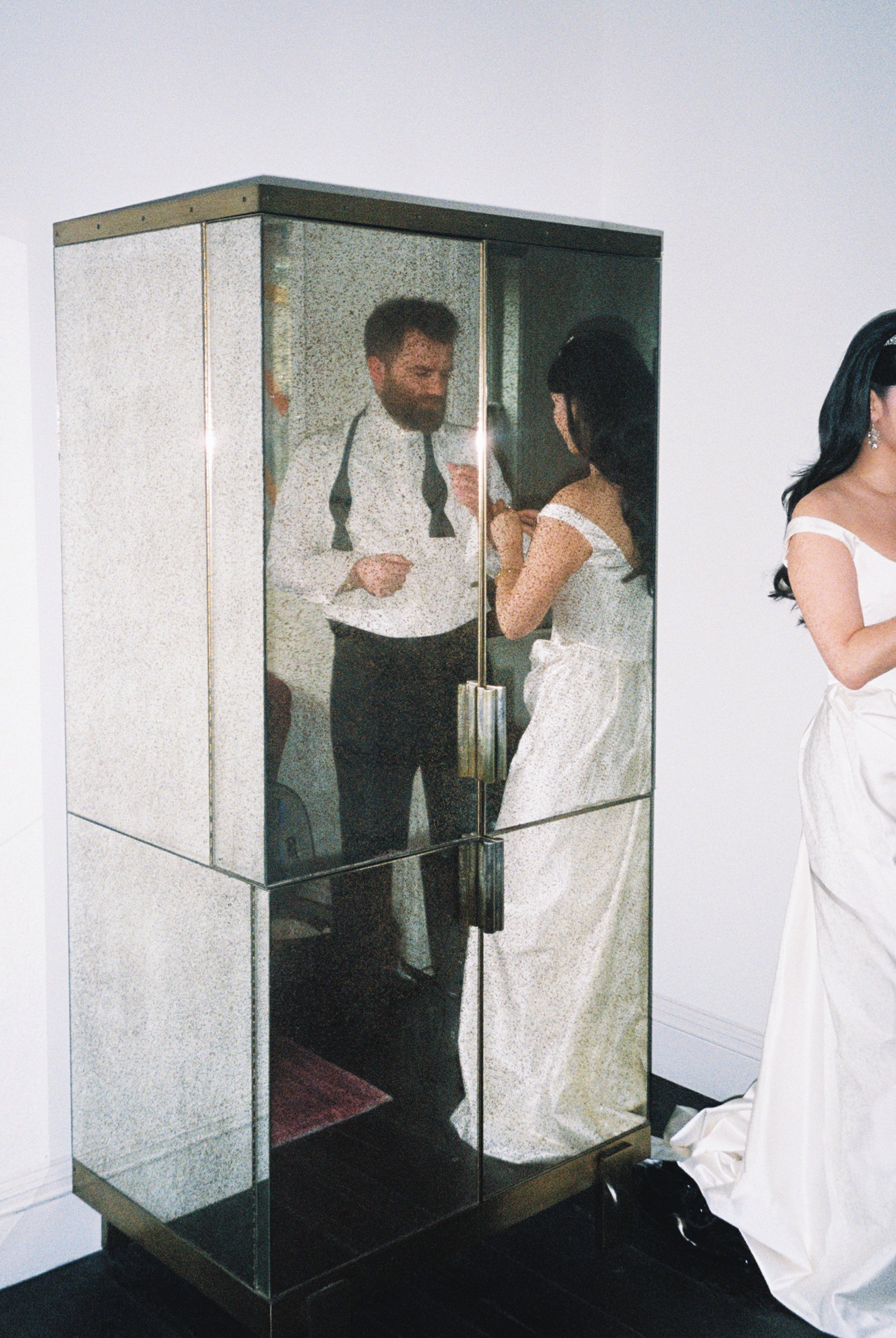 A glass and metal vending machine or display case with a person inside, appearing to be a man with a beard, wearing a white shirt and black tie, seen through the reflection. Another person in a white dress is standing nearby, with part of her face an