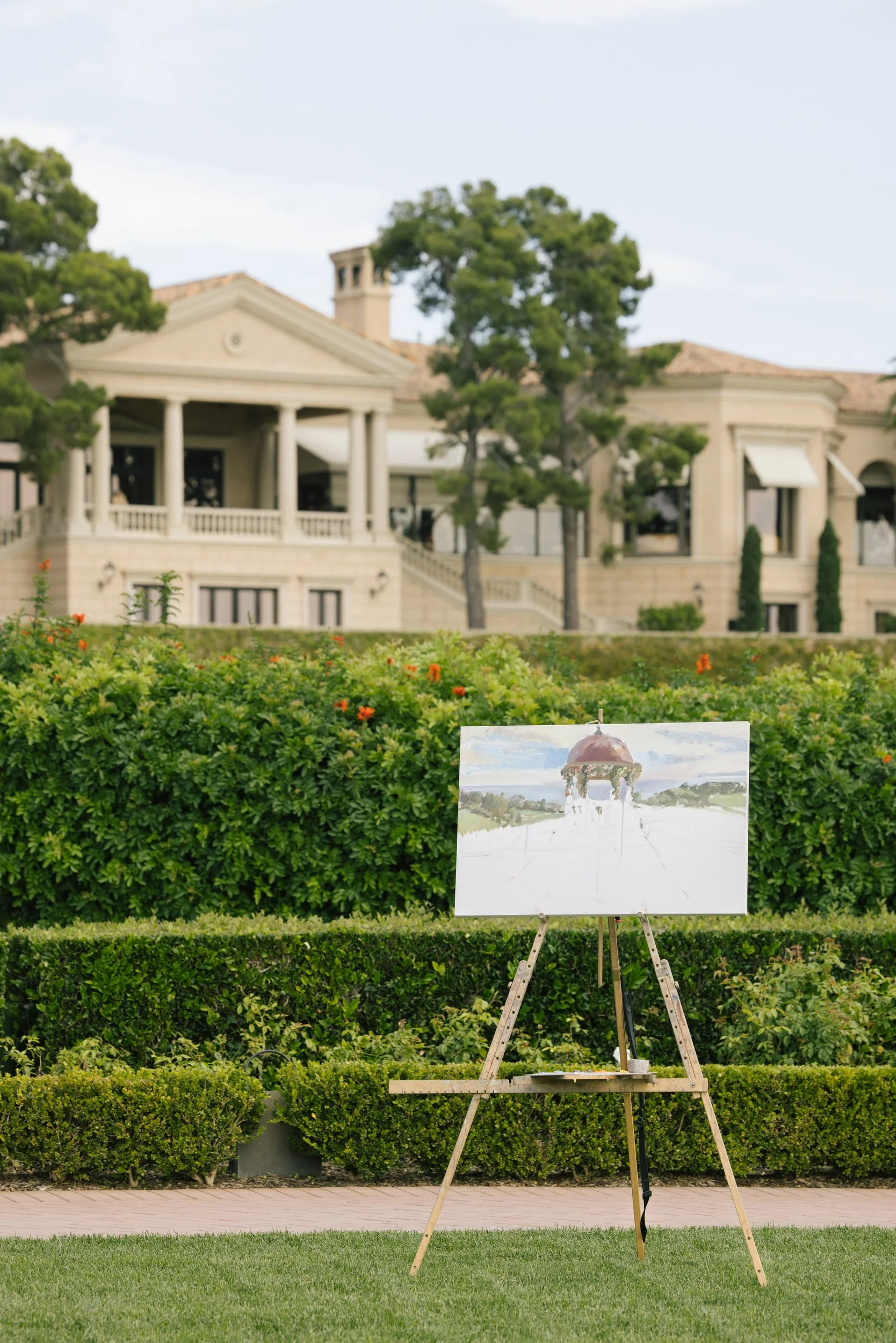 An outdoor scene with a painting on an easel in the foreground, in front of a neatly trimmed hedge and green grass. Behind are trees and a large, elegant house with a porch, columns, and a chimney, under a partly cloudy sky.