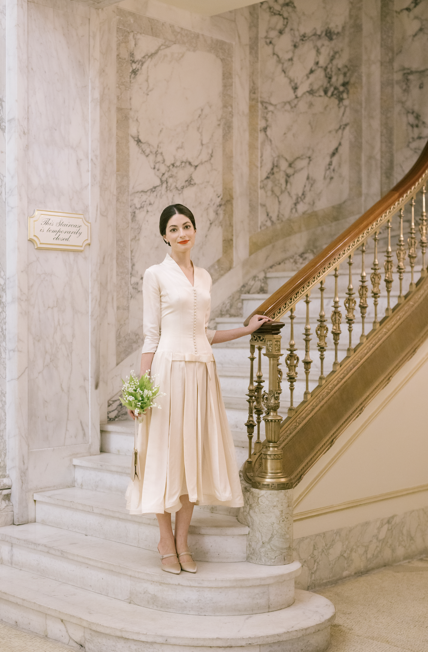 A woman in a vintage cream-colored dress holding a bouquet of white and green flowers is standing on marble stairs in a grand interior with marble walls and a curved staircase with a wooden banister.