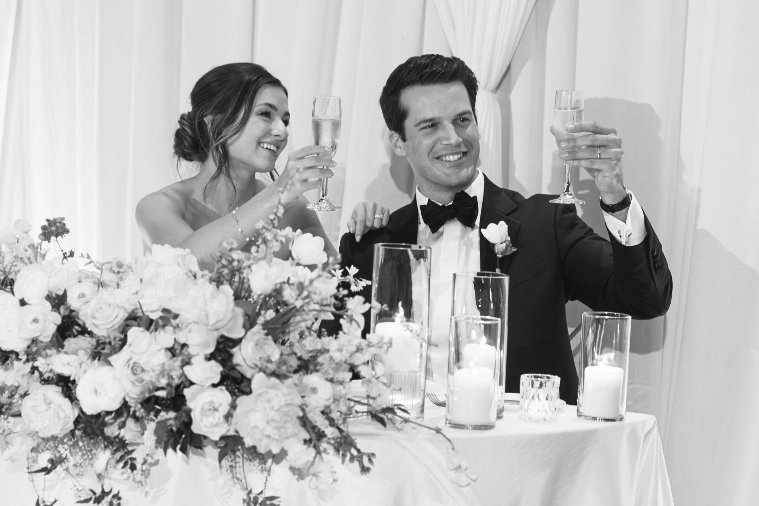 A black and white photo of a newlywed couple at their wedding reception. The bride and groom are sitting together, smiling, holding champagne glasses for a toast. The table is decorated with a large bouquet of flowers and several candles.