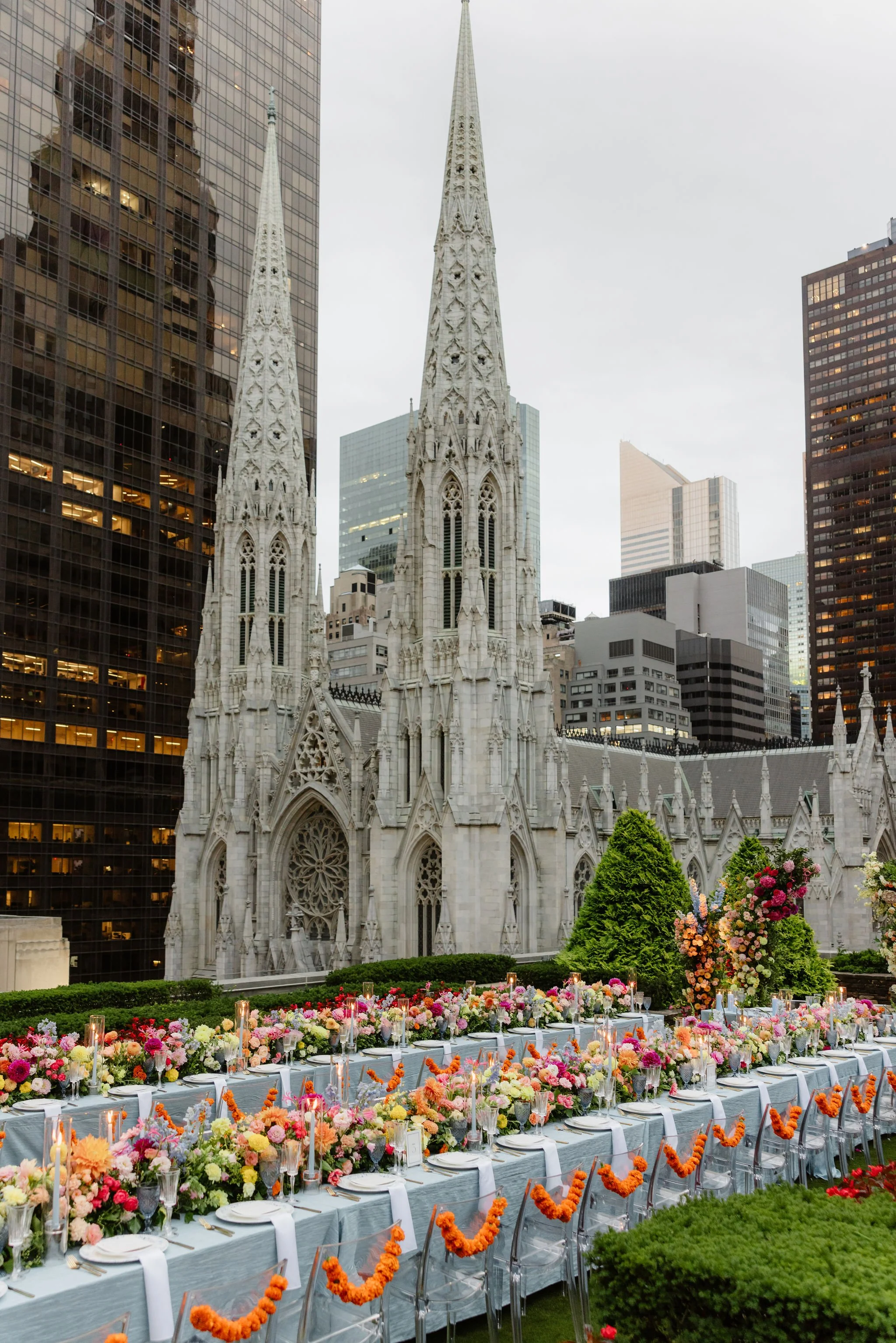 A wedding reception setup with long banquet tables decorated with colorful flowers, candles, and white tablecloths, set outdoors in front of a historic white church with pointed steeples, surrounded by tall modern skyscrapers.