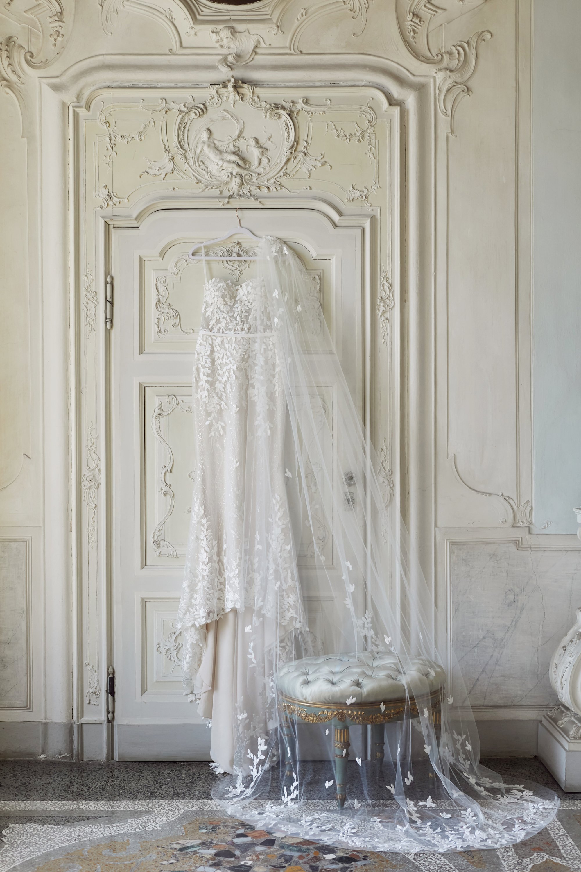 A white lace wedding dress hanging on a hanger with a long veil, displayed against a decorated cream-colored wall with intricate molding and a small upholstered stool underneath.