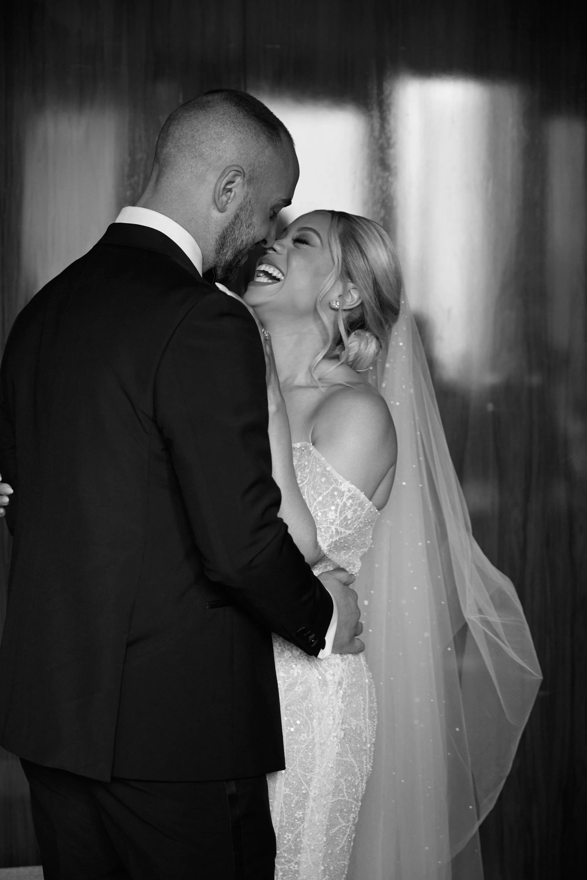 A black and white photo of a smiling bride in a wedding dress and veil being close to a groom in a tuxedo, with their foreheads touching.