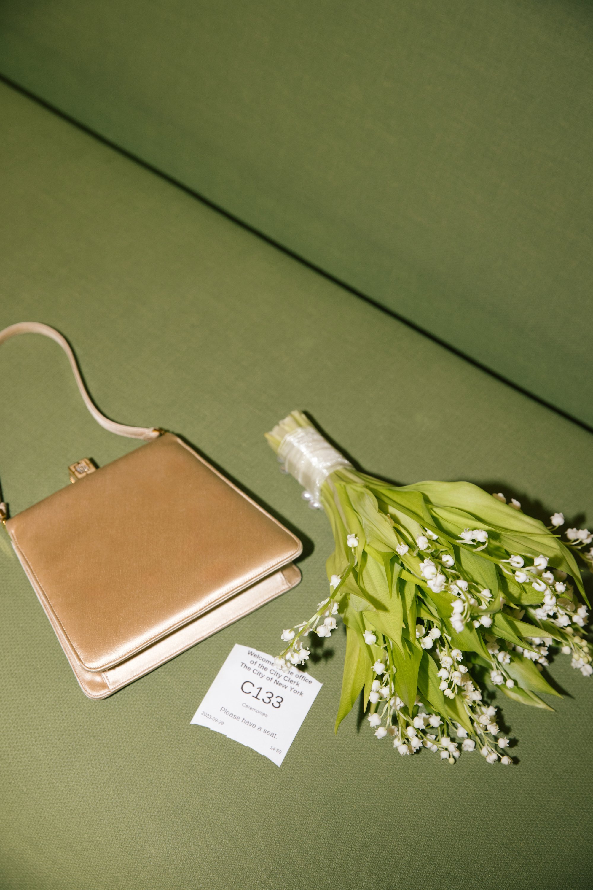 A bouquet of white flowers with green leaves resting on a green surface, next to a gold-colored purse and a white card with black text. The card reads 'Welcome to the office of the City Clerk, The City of New York, C133, Ceremonies, Please have a sea