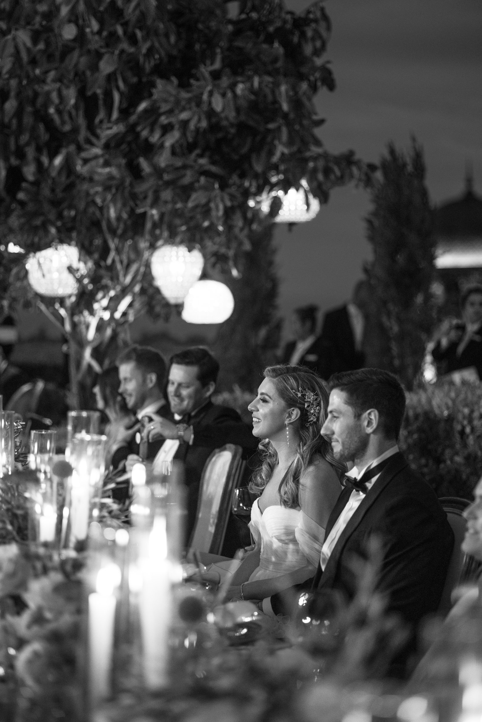Black and white photograph of a wedding reception. The bride and groom are seated at a long table with guests, under hanging lanterns and surrounded by trees.