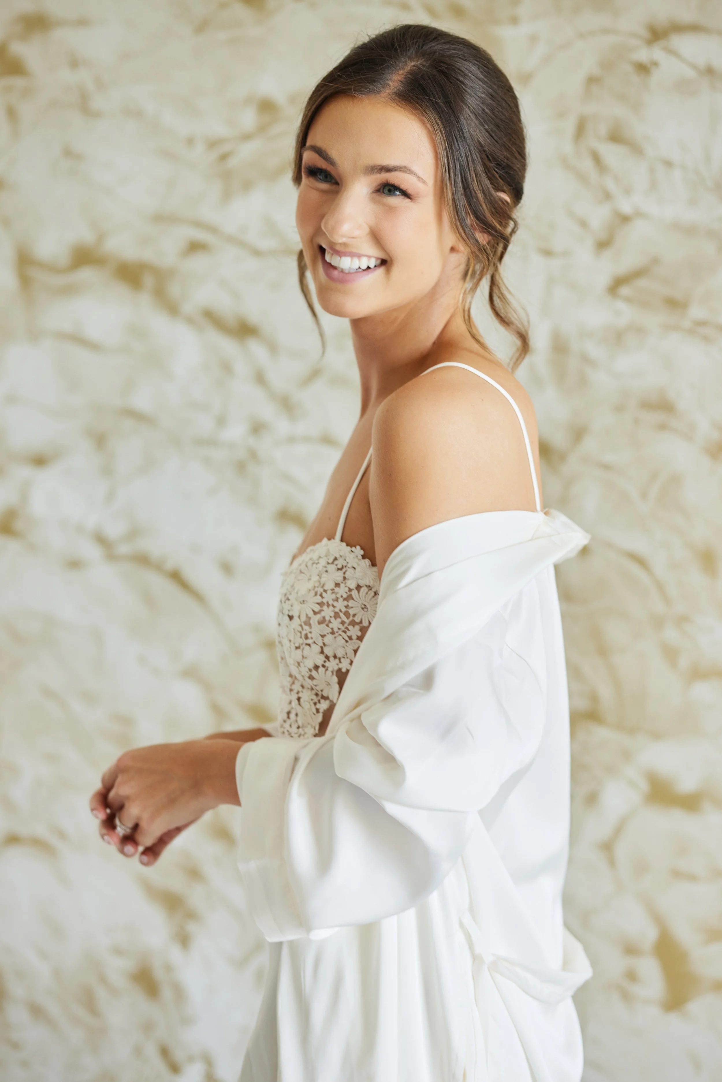 A young woman with brown hair styled in loose waves, wearing a white lace dress and a white satin robe off the shoulder, smiling brightly with a blurred light-colored stone wall in the background.