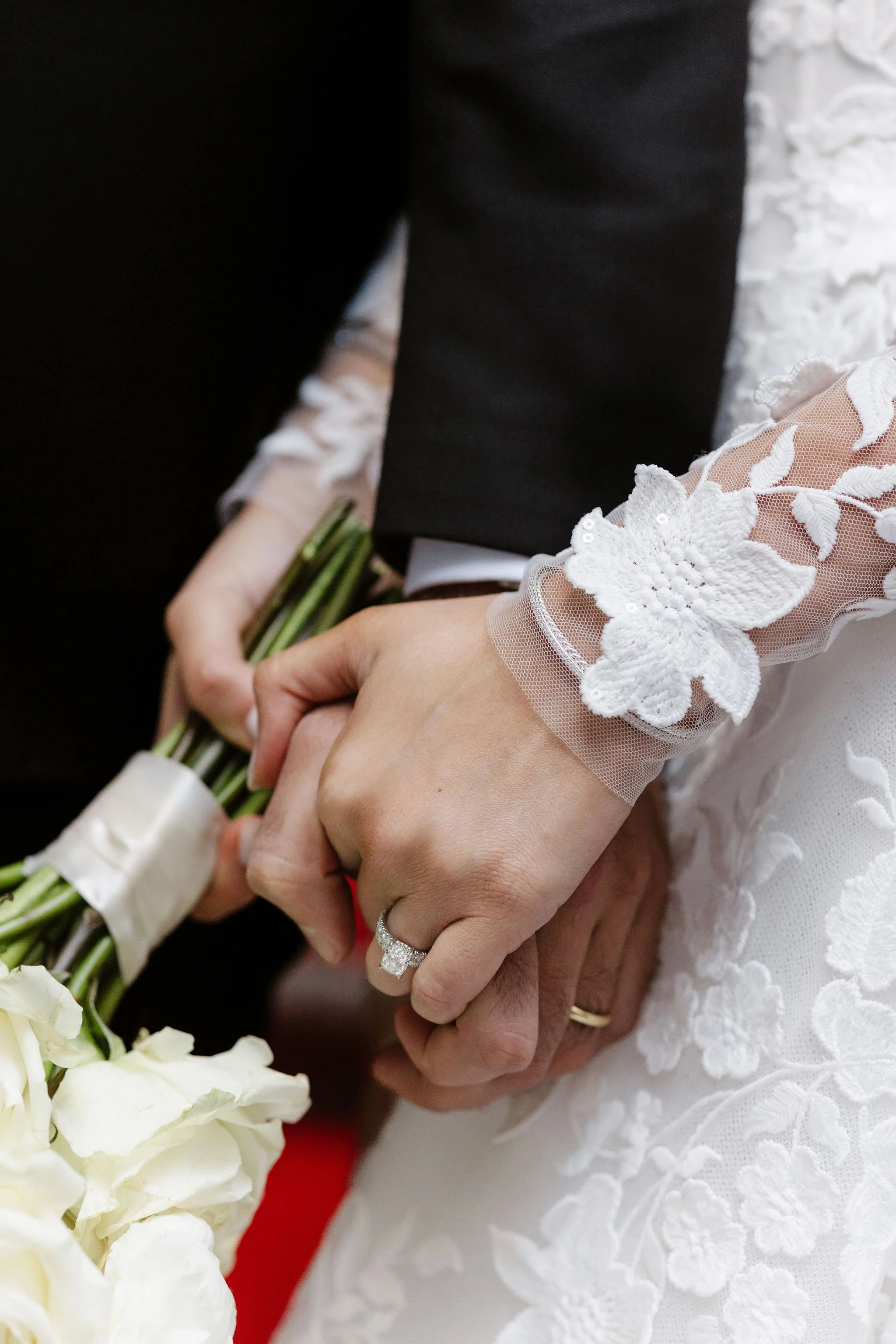 Close-up of a bride and groom holding hands, the bride wearing a white lace gown with floral embellishments and a wedding ring, and the groom in a black suit, with a bouquet of white flowers in the background.